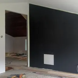 Ciemna ściana po szpachlowaniu i malowaniu, widoczna instalacja elektryczna i fragment sufitu pod skosem. W tle widoczne przygotowania do dalszych prac wykończeniowych.