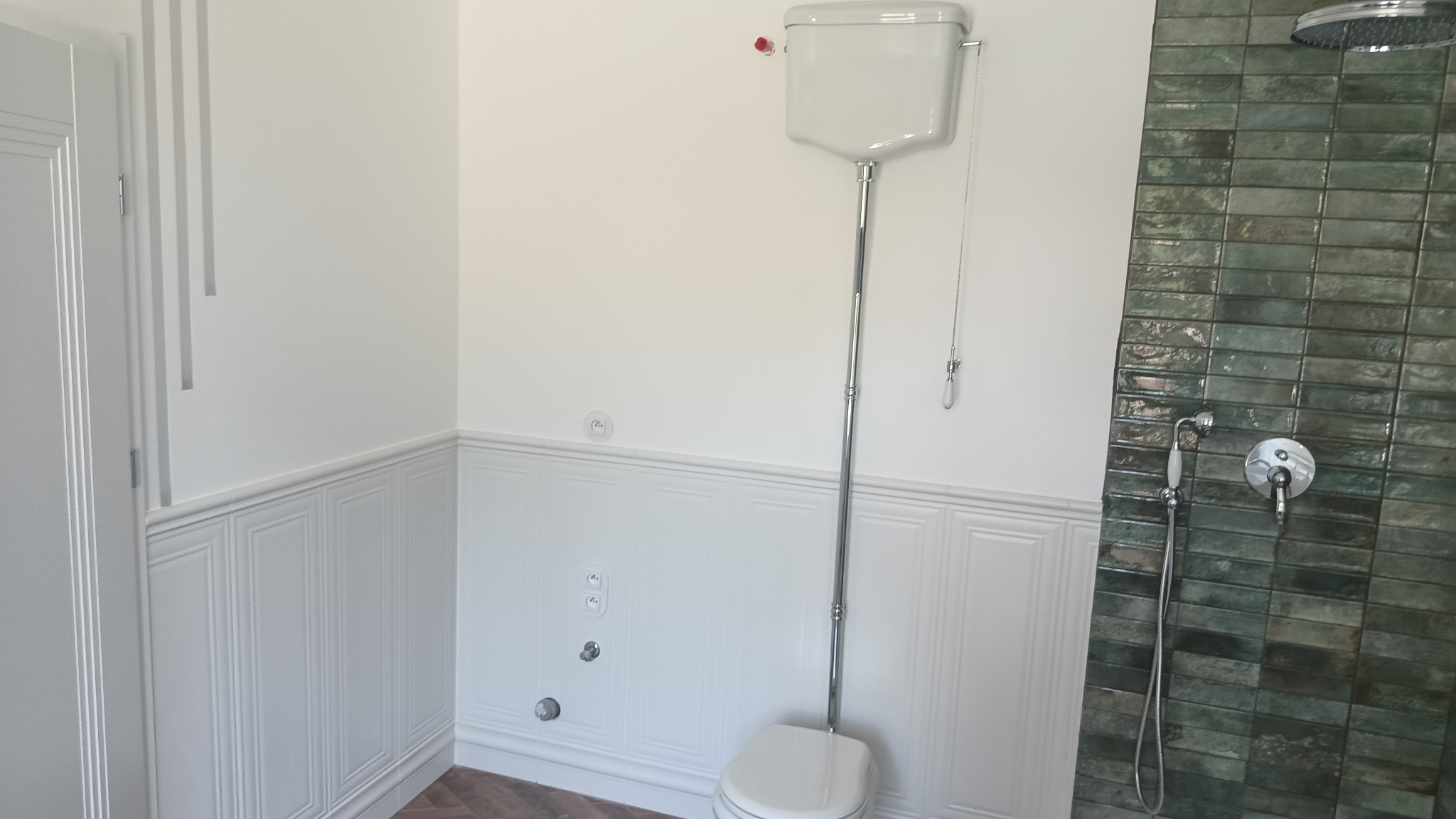 Biała łazienka z panelami ściennymi, tradycyjnym WC z wysokim zbiornikiem i zieloną ścianą prysznicową z płytek. Widoczne detale wykończeniowe.