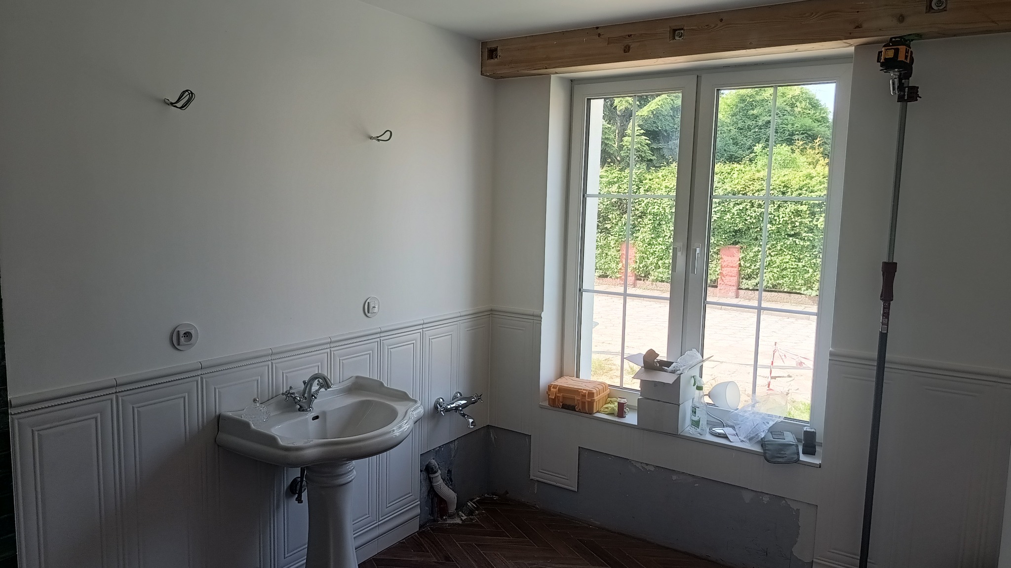 Remont łazienki w stylu retro: biała umywalka na postumencie, boazeria, częściowo przygotowana ściana pod zabudowę. Widoczny laser krzyżowy.