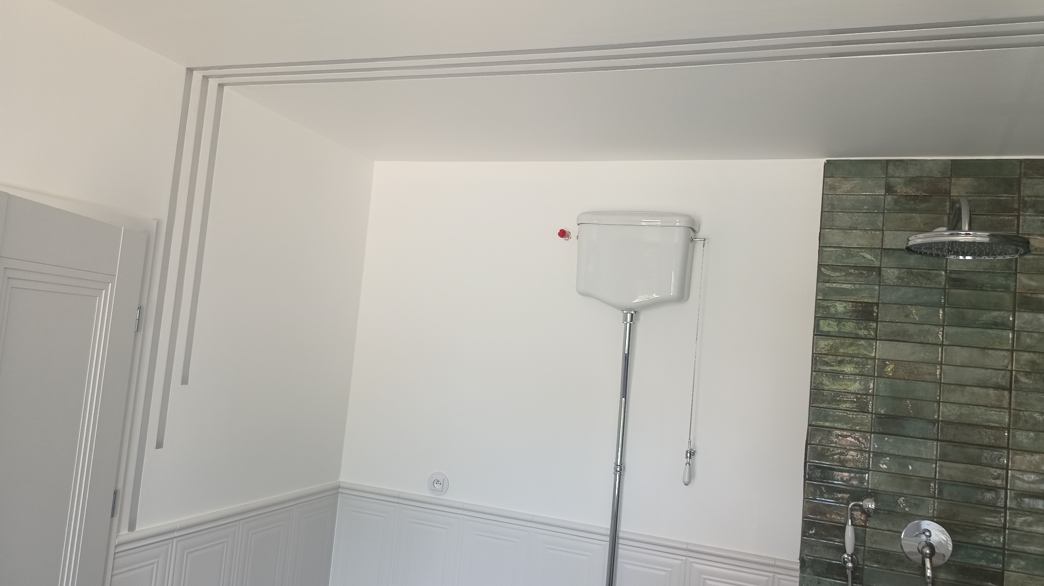 Wykończona łazienka: białe ściany z dekoracyjnymi listwami, toaleta ze spłuczką na ścianie i zielona ściana z płytek z deszczownicą. Minimalistyczny design.