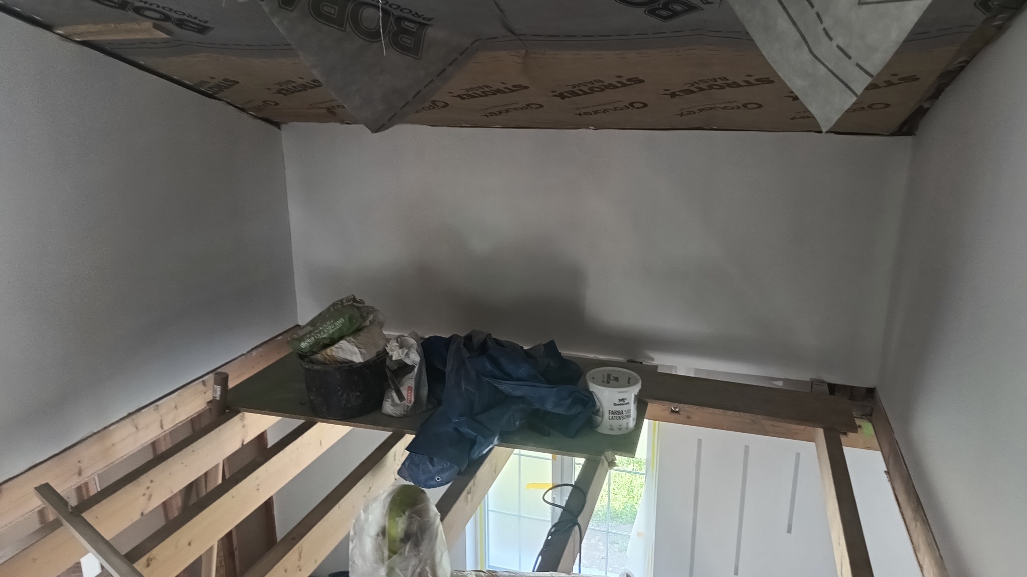 Wykończone poddasze ze ścianami pomalowanymi na biało. Widoczne materiały budowlane na drewnianym podeście, okno w tle. Ukończony etap prac.