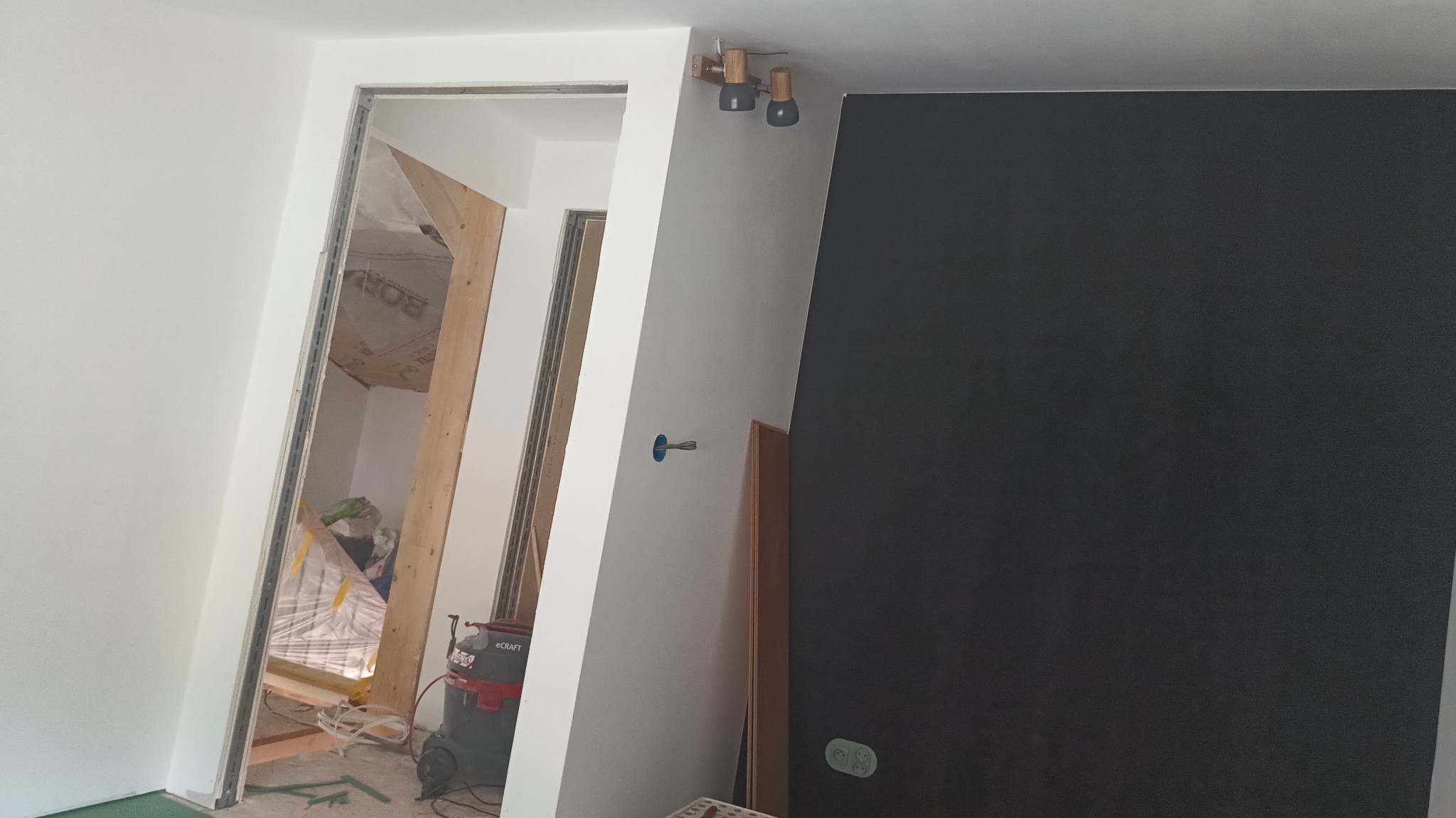 Montaż ścianki działowej: widoczny profil, izolacja, odkurzacz budowlany. Częściowo wykończona ściana w kolorze grafitowym z gniazdkiem elektrycznym.