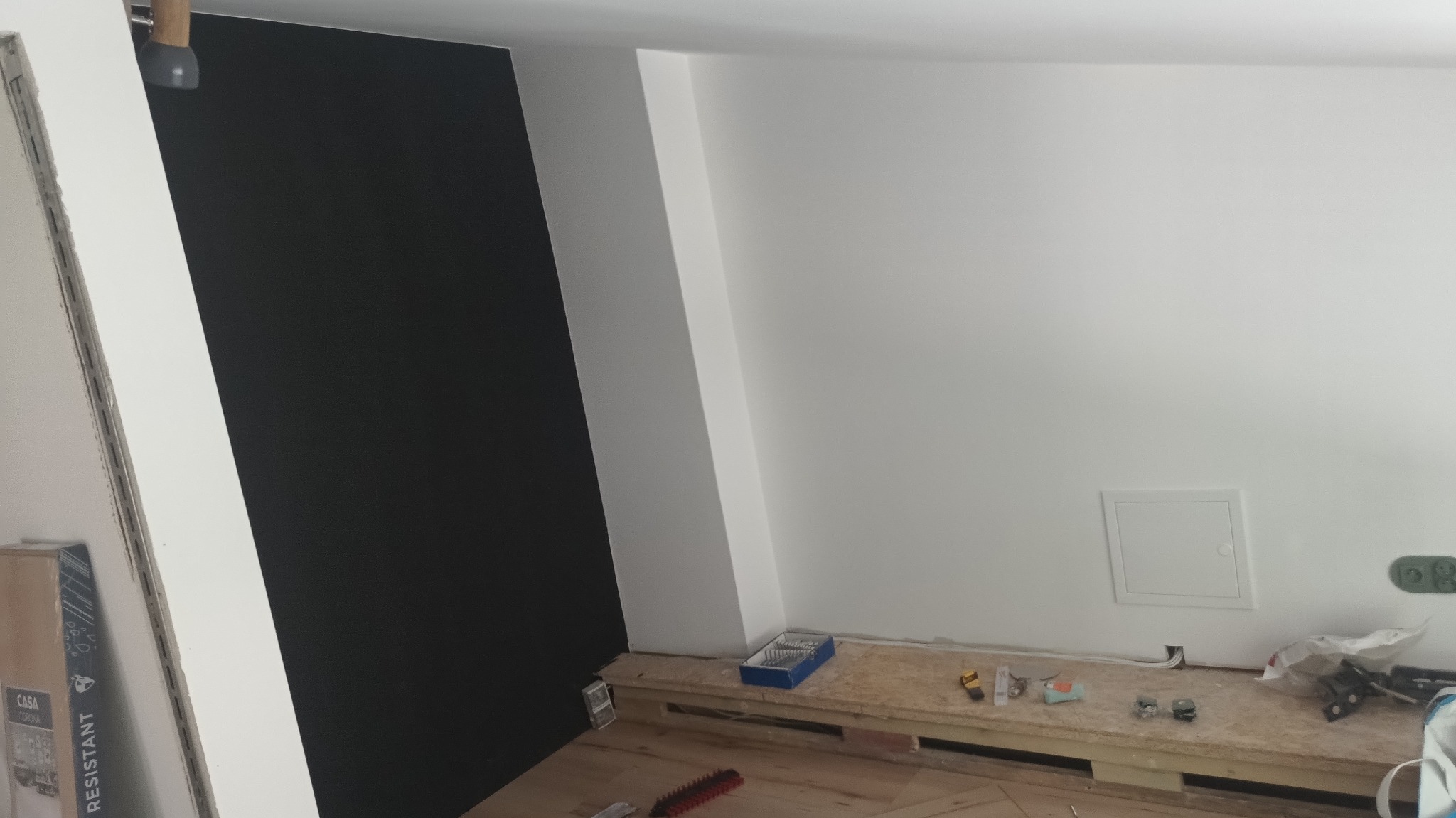 Ściana po montażu płyt w kolorze grafitowym, sąsiadująca z białą ścianą. Na podłodze widoczne narzędzia i fragmenty materiałów budowlanych, co wskazuje na etap wykończeniowy.