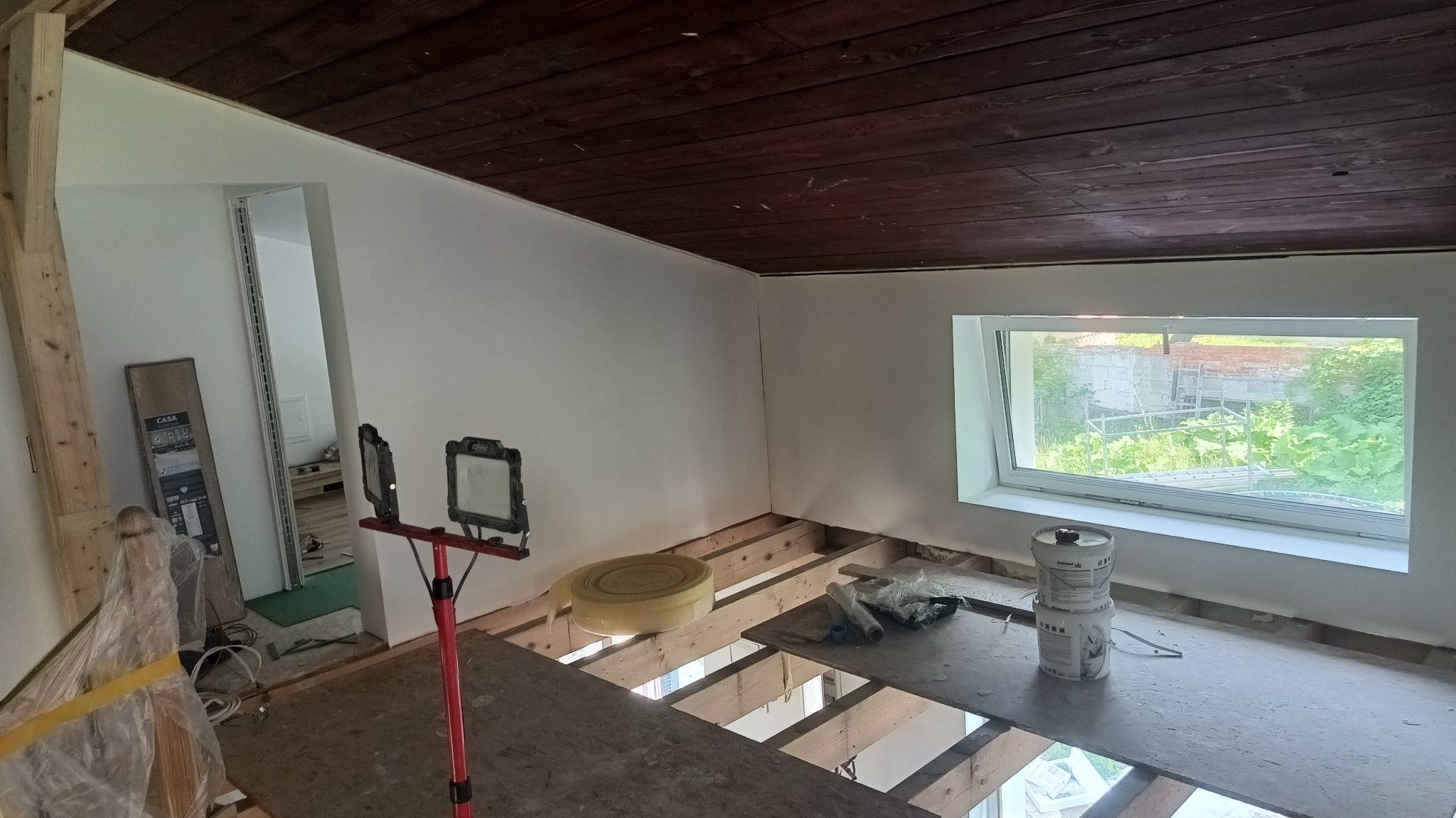 Wnętrze w trakcie prac wykończeniowych: sufit z ciemnego drewna, białe ściany, okno z widokiem na zieleń. Na podłodze materiały budowlane i narzędzia.
