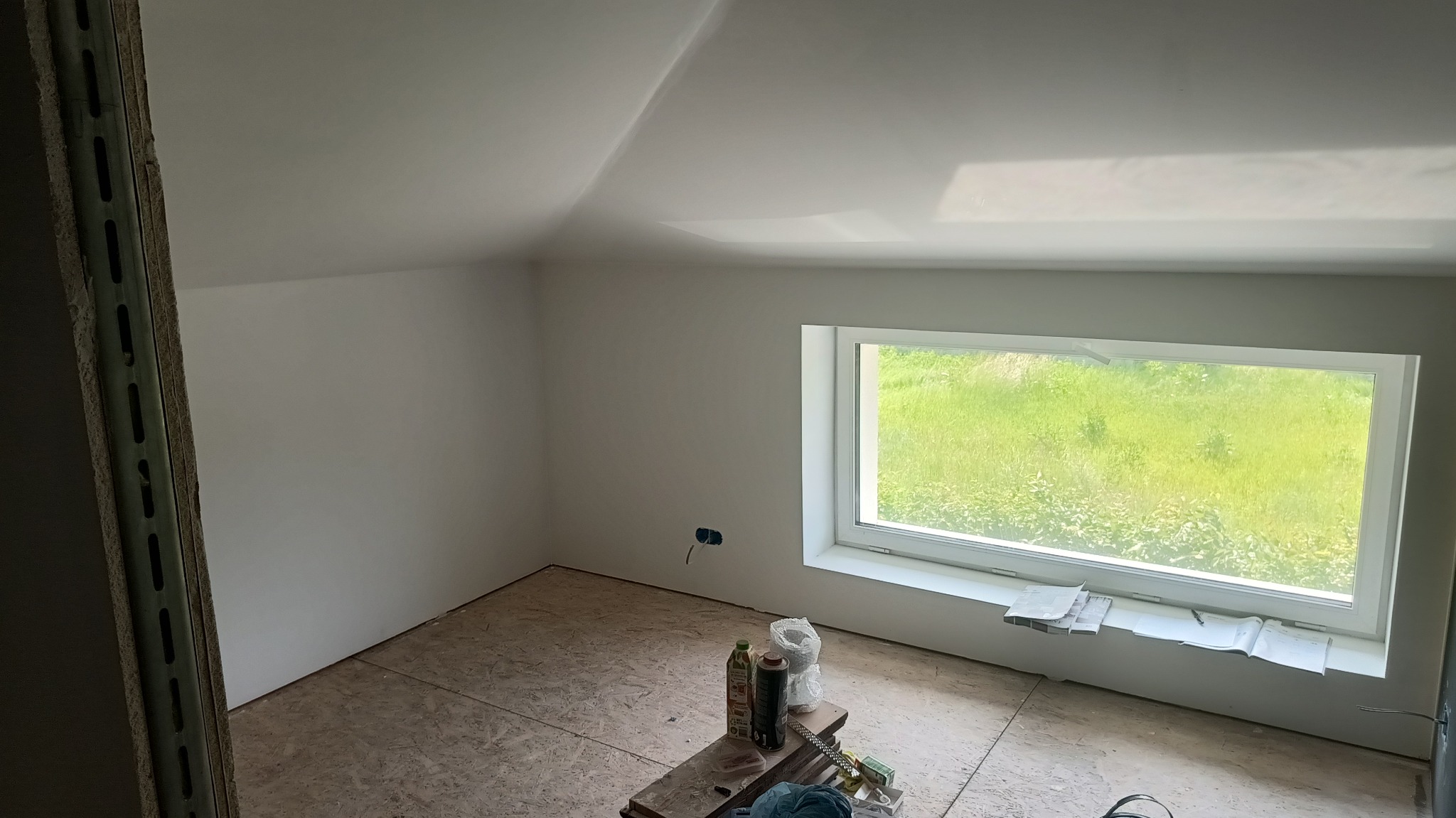 Poddasze po pracach wykończeniowych: białe ściany i sufit, okno z widokiem na zieleń, podłoga z płyt OSB, widoczne narzędzia i materiały malarskie na podeście.