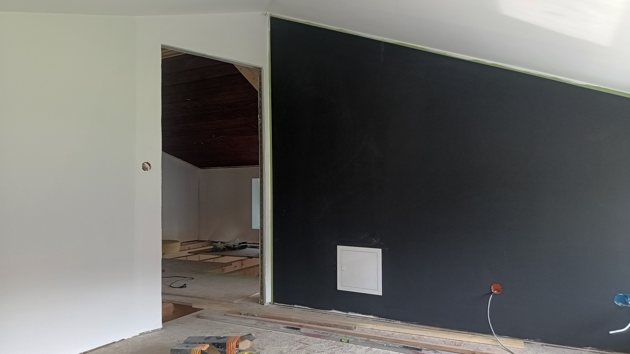 Ciemna ściana po szpachlowaniu i malowaniu, widoczna instalacja elektryczna i fragment sufitu pod skosem. W tle widoczne przygotowania do dalszych prac wykończeniowych.