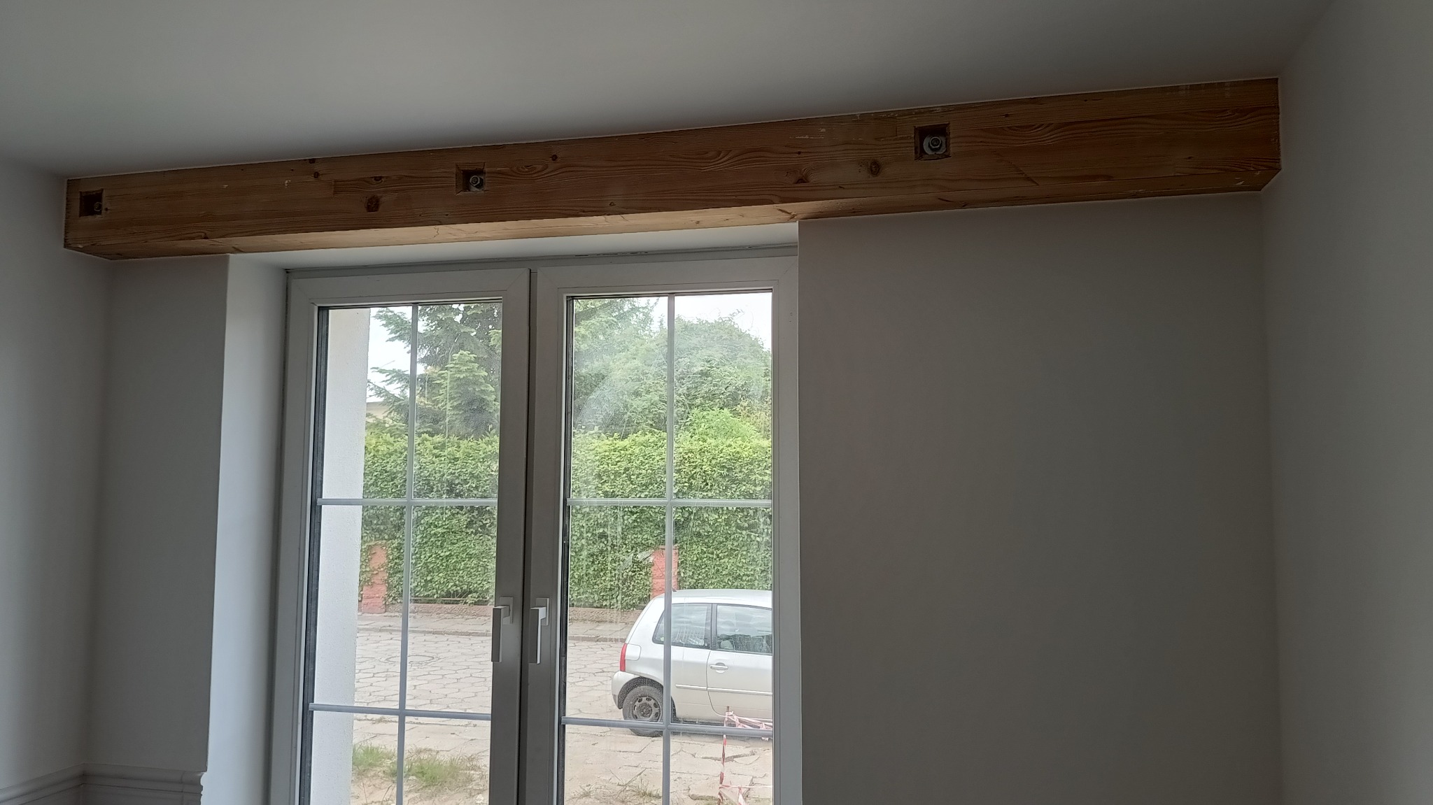Drewniana belka dekoracyjna nad oknem w jasnym wnętrzu. Widoczne otwory na oświetlenie. Za oknem zieleń i zaparkowany samochód.