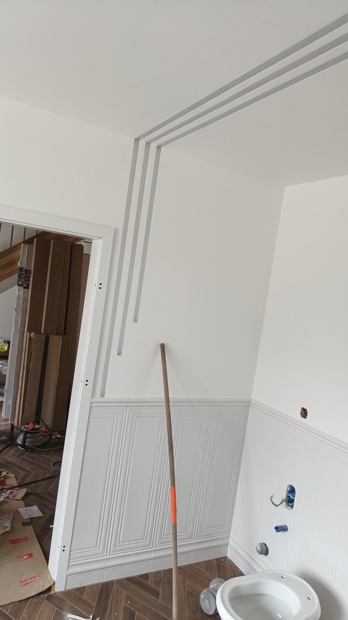 Wykończone wnętrze łazienki z dekoracyjnymi panelami ściennymi, listwami sufitowymi i częściowo zamontowaną armaturą. Widoczny fragment podłogi z parkietem.