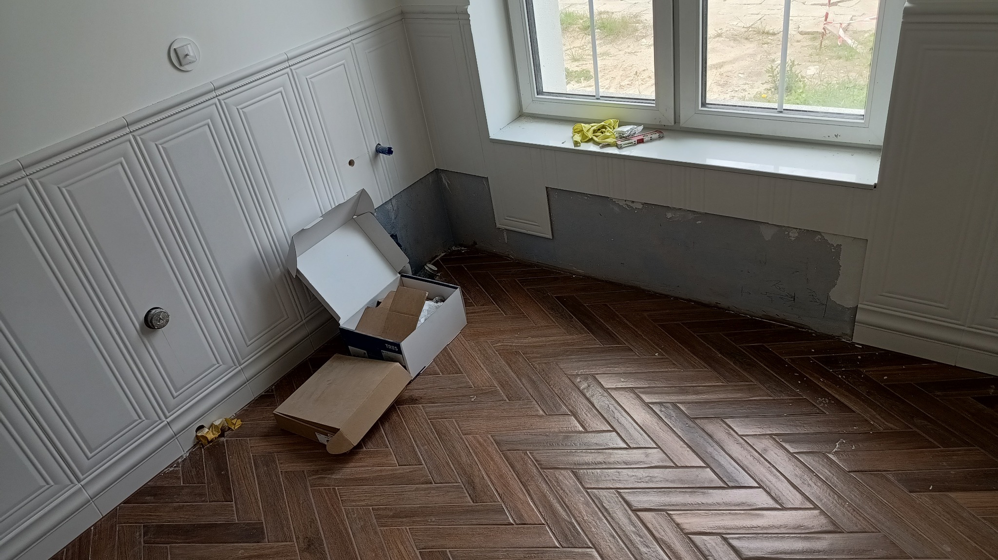 Wnętrze w trakcie prac wykończeniowych: ściana z dekoracyjną boazerią, fragment ściany przygotowany pod montaż, podłoga w jodełkę i otwarte pudełko z materiałami.