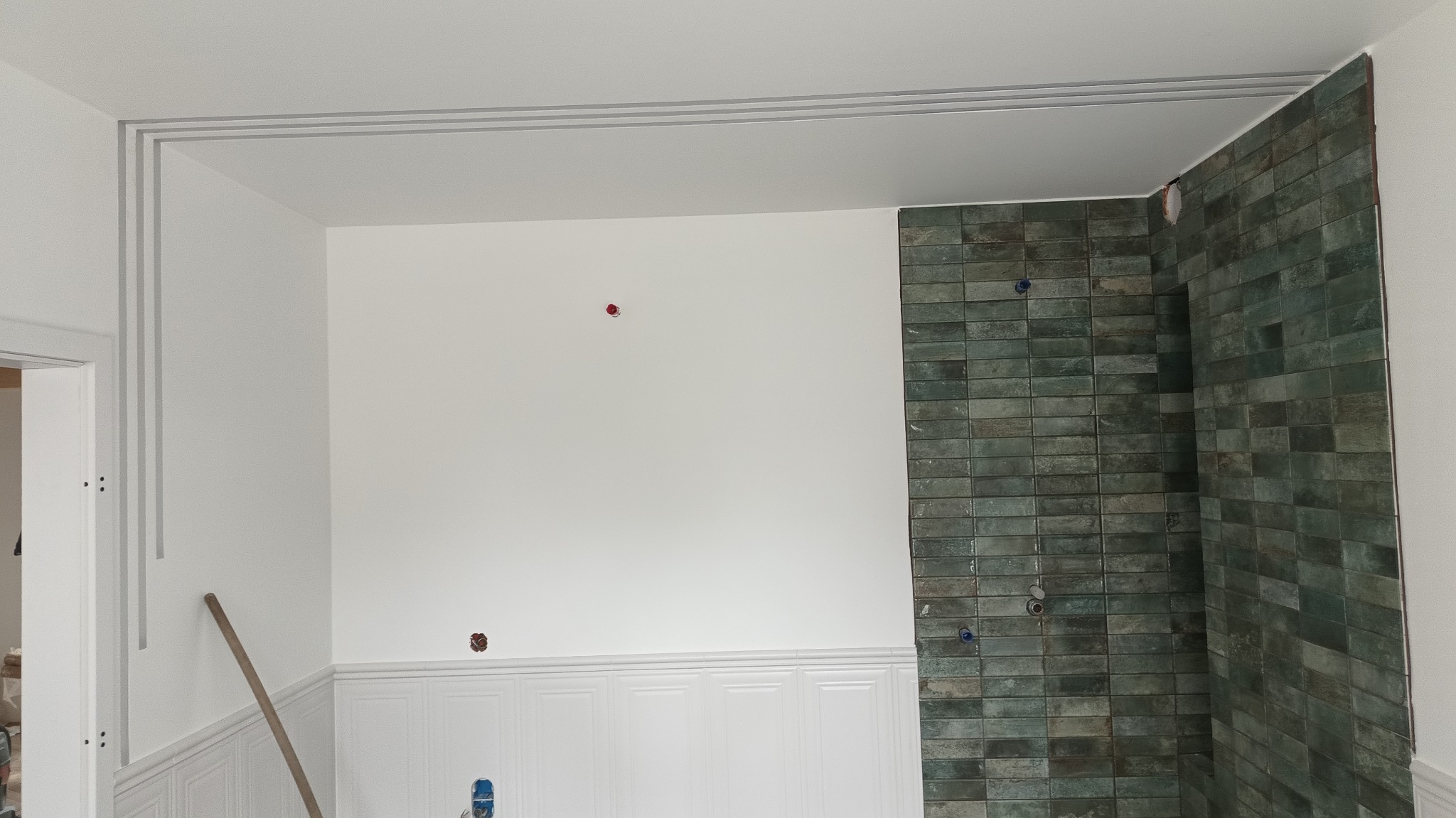 Wnętrze w trakcie remontu: białe ściany z dekoracyjną listwą, częściowo położone zielone płytki na ścianie, widoczne punkty instalacji elektrycznej i wodnej.