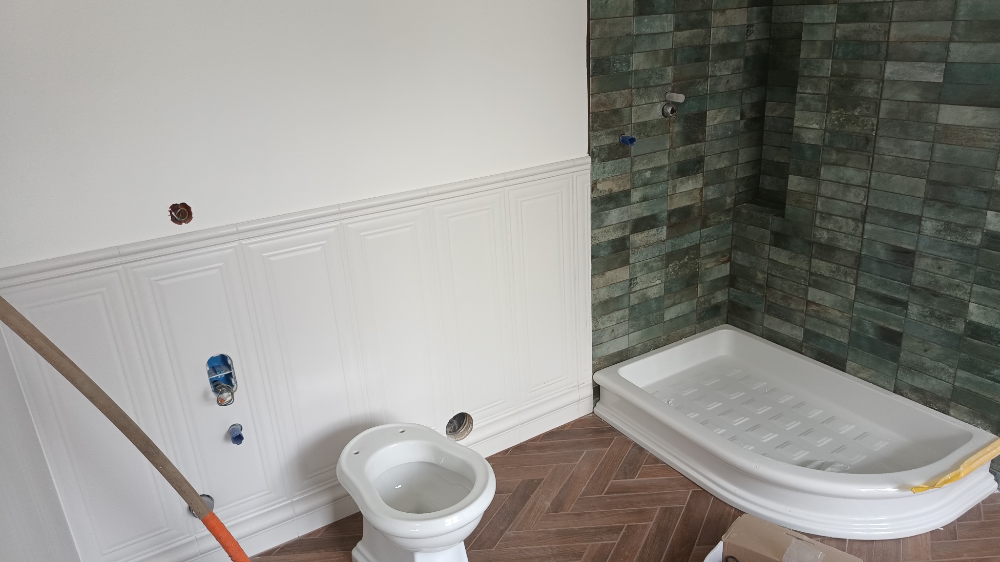 Remont łazienki w trakcie: biała boazeria, zielona glazura, brązowa podłoga w jodełkę, biały brodzik i bidet, widoczne instalacje. Prace wykończeniowe.