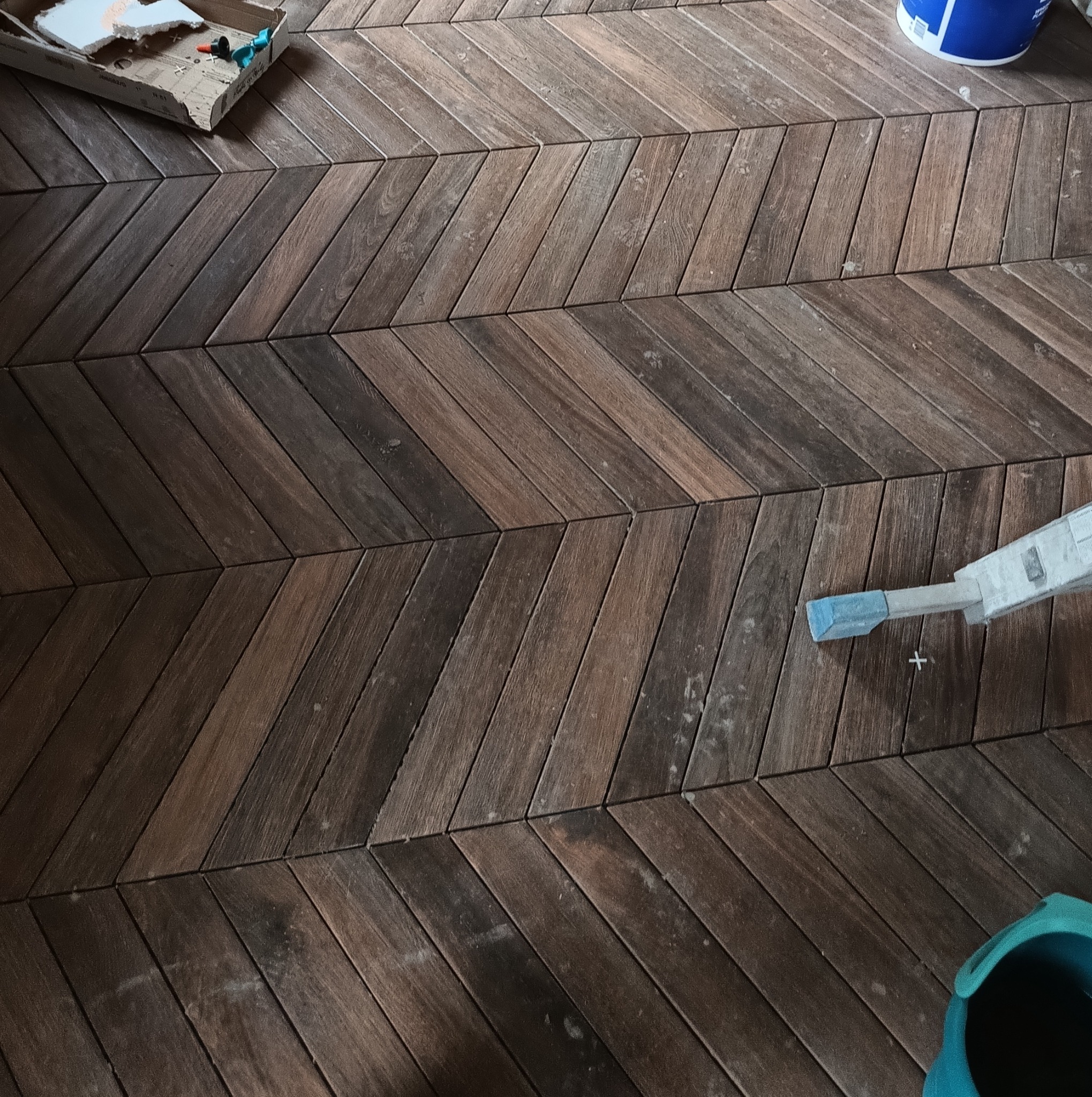 Układanie parkietu w jodełkę z ciemnego drewna. Widoczne akcesoria montażowe, w tym krzyżyki dystansowe, karton i wiaderko z klejem.