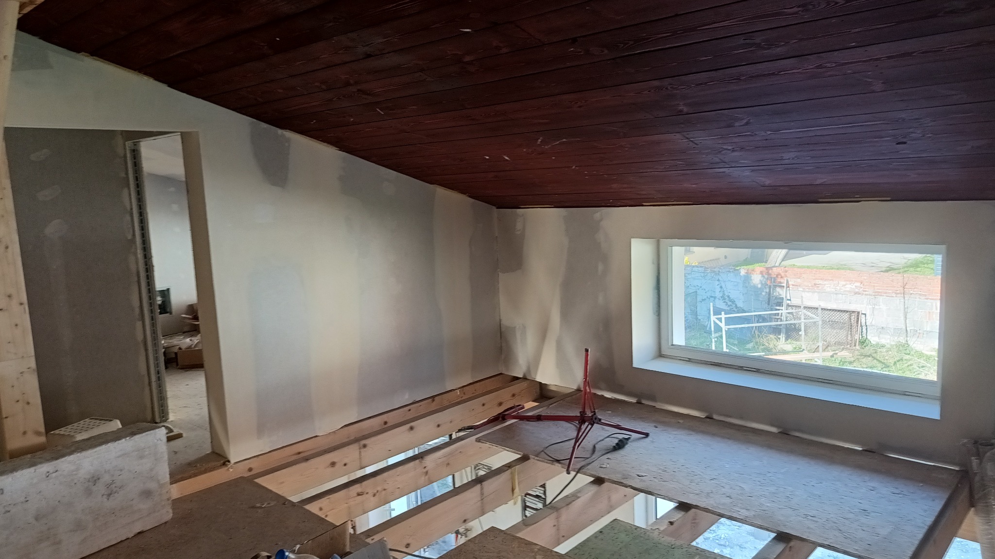 Poddasze w trakcie wykańczania: ściany przygotowane do gładzi, widoczne łączenia płyt. Ciemne drewniane deski na suficie i okno z widokiem na zewnątrz.