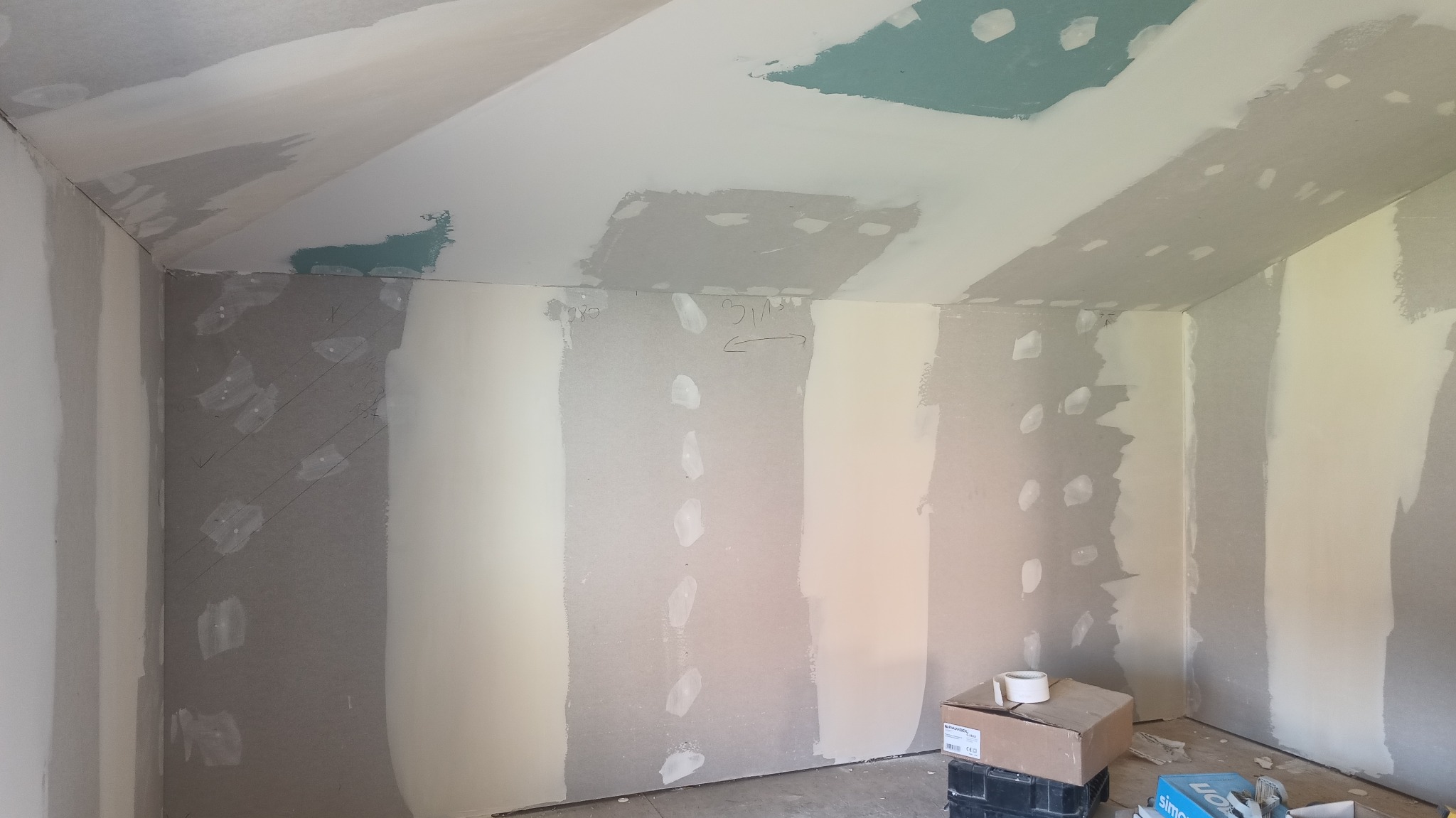 Wnętrze pokoju w trakcie prac wykończeniowych ścian. Widoczne szpachlowane łączenia płyt, zaznaczone linie cięć i narzędzia na podłodze, co sugeruje etap przygotowania do malowania.