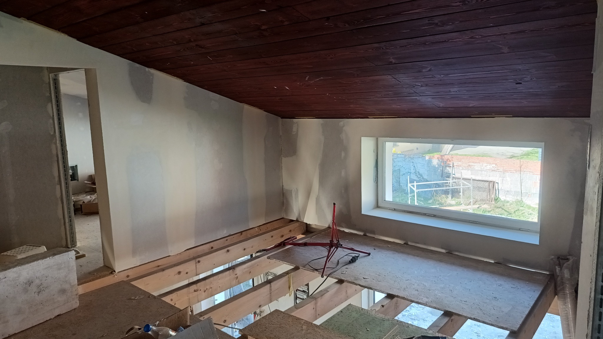 Poddasze w trakcie adaptacji: ściany przygotowane pod wykończenie, widoczne drewniane legary pod podłogą. Ciemna, drewniana boazeria na suficie.