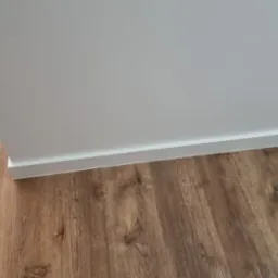 Ściana pomalowana na biało z zamontowaną białą listwą przypodłogową i podłogą z paneli imitujących drewno w ciepłym odcieniu brązu, fragment narożnika pokoju.