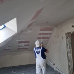 Mariusz Szczepański M M Wykończenia Wnętrz - Pracownik w białym kombinezonie aplikuje szpachlę natryskową na skośny sufit poddasza. Widoczne łatki szpachli i okno dachowe z niebieską taśmą.