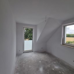 Mariusz Szczepański M M Wykończenia Wnętrz - Wnętrze pokoju na poddaszu po szpachlowaniu i malowaniu na biało. Widoczne okno z widokiem na zieleń i pole, surowa podłoga, gładkie ściany. Przygotowanie do dalszych prac wykończeniowych.