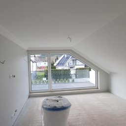 Mariusz Szczepański M M Wykończenia Wnętrz - Pokój na poddaszu po malowaniu na biało, z oknem dachowym i listwami ozdobnymi. Widoczne przewody elektryczne i zabezpieczony materiał budowlany.