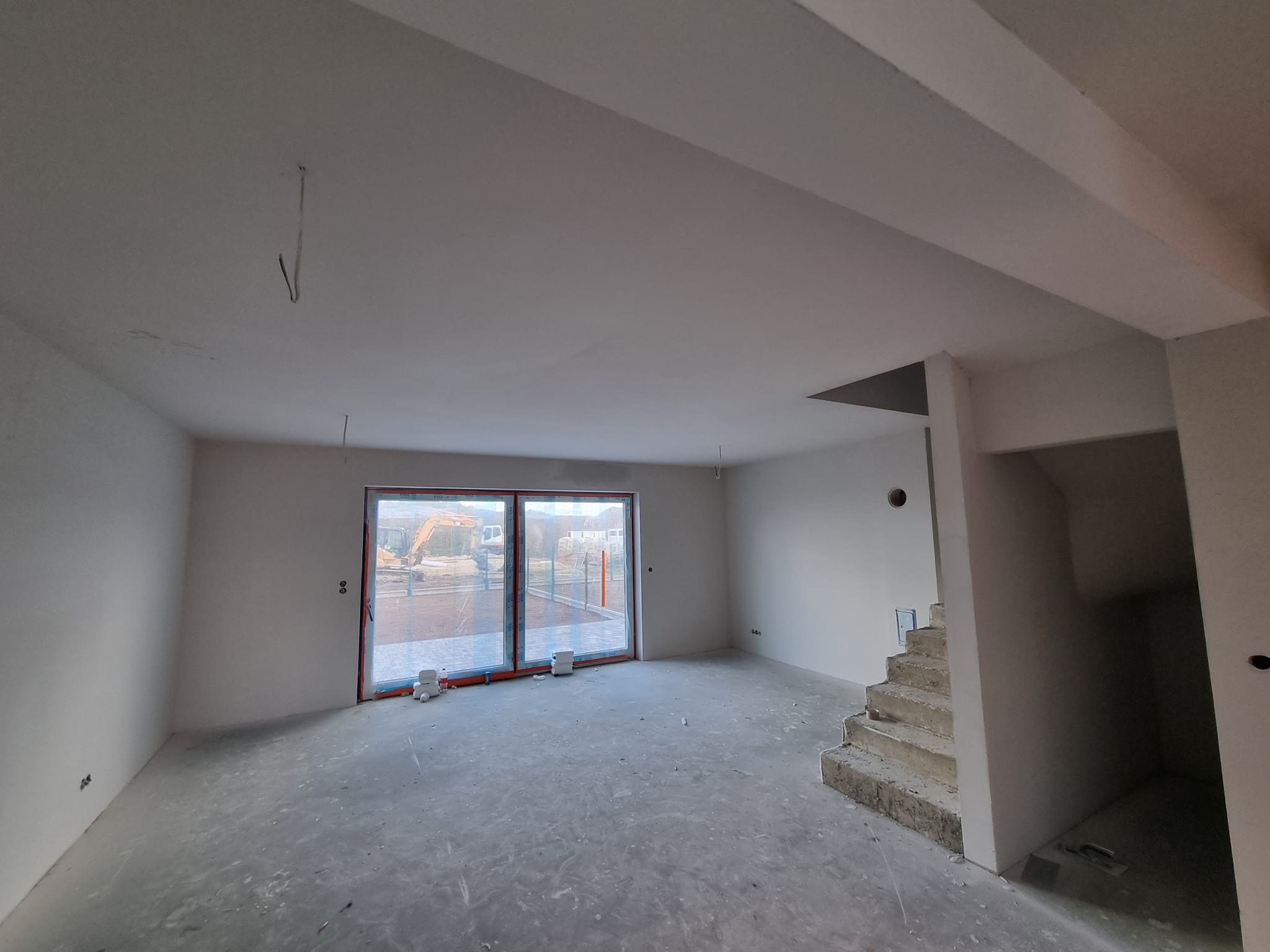 Wnętrze w stanie deweloperskim: surowe ściany po szpachlowaniu, betonowa posadzka, widoczne schody i okno z widokiem na plac budowy z koparką.