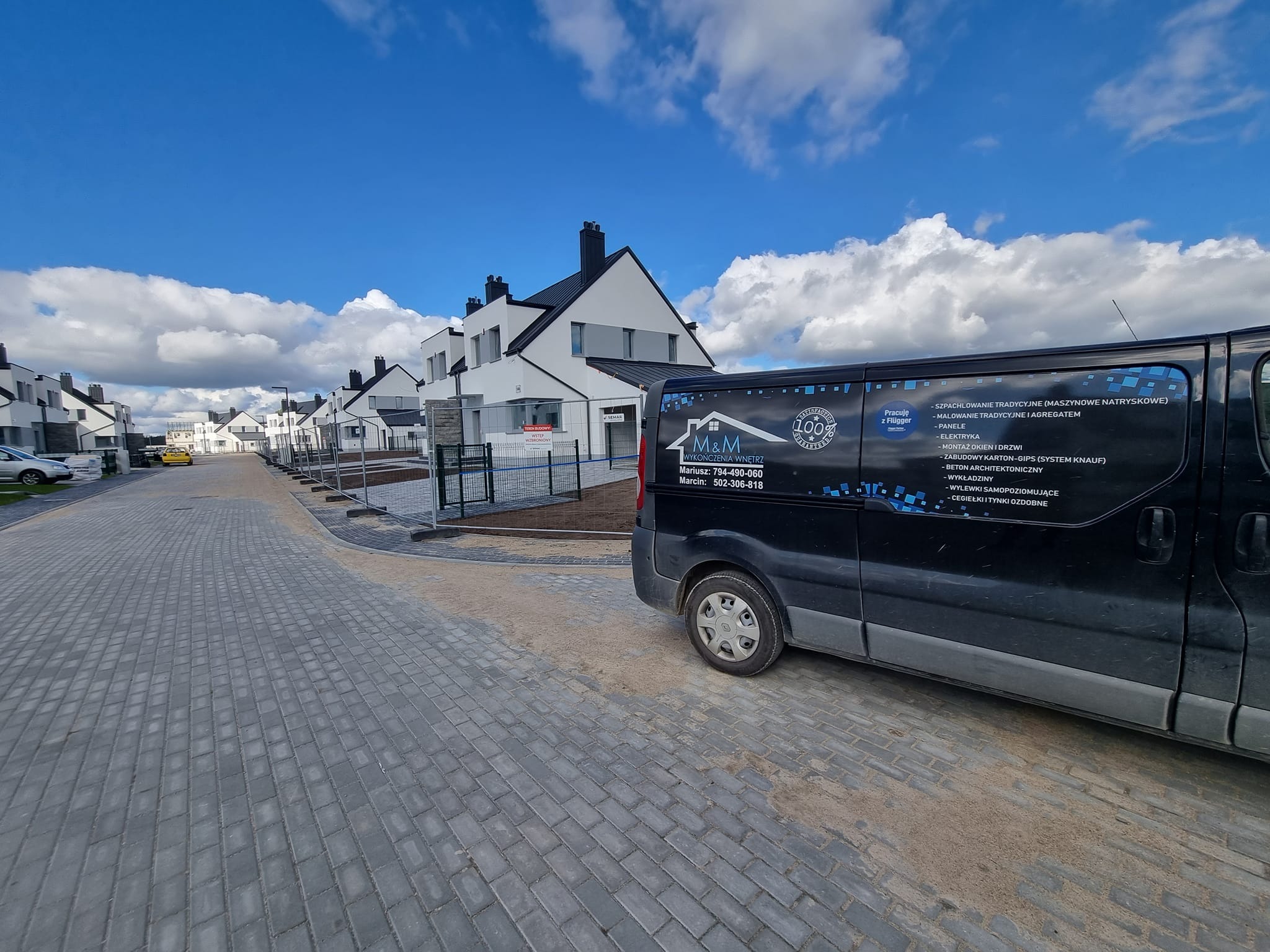 Czarny van firmy M&M Wykończenia Wnętrz z ofertą usług (szpachlowanie, malowanie, panele, elektryka) na tle osiedla nowych domów w słoneczny dzień.