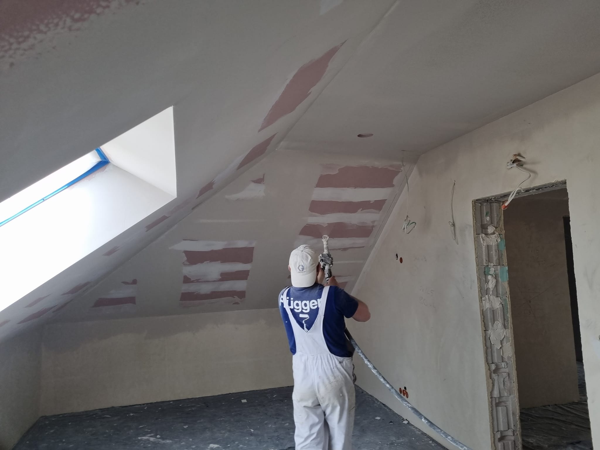 Pracownik w białym kombinezonie aplikuje szpachlę natryskową na skośny sufit poddasza. Widoczne łatki szpachli i okno dachowe z niebieską taśmą.