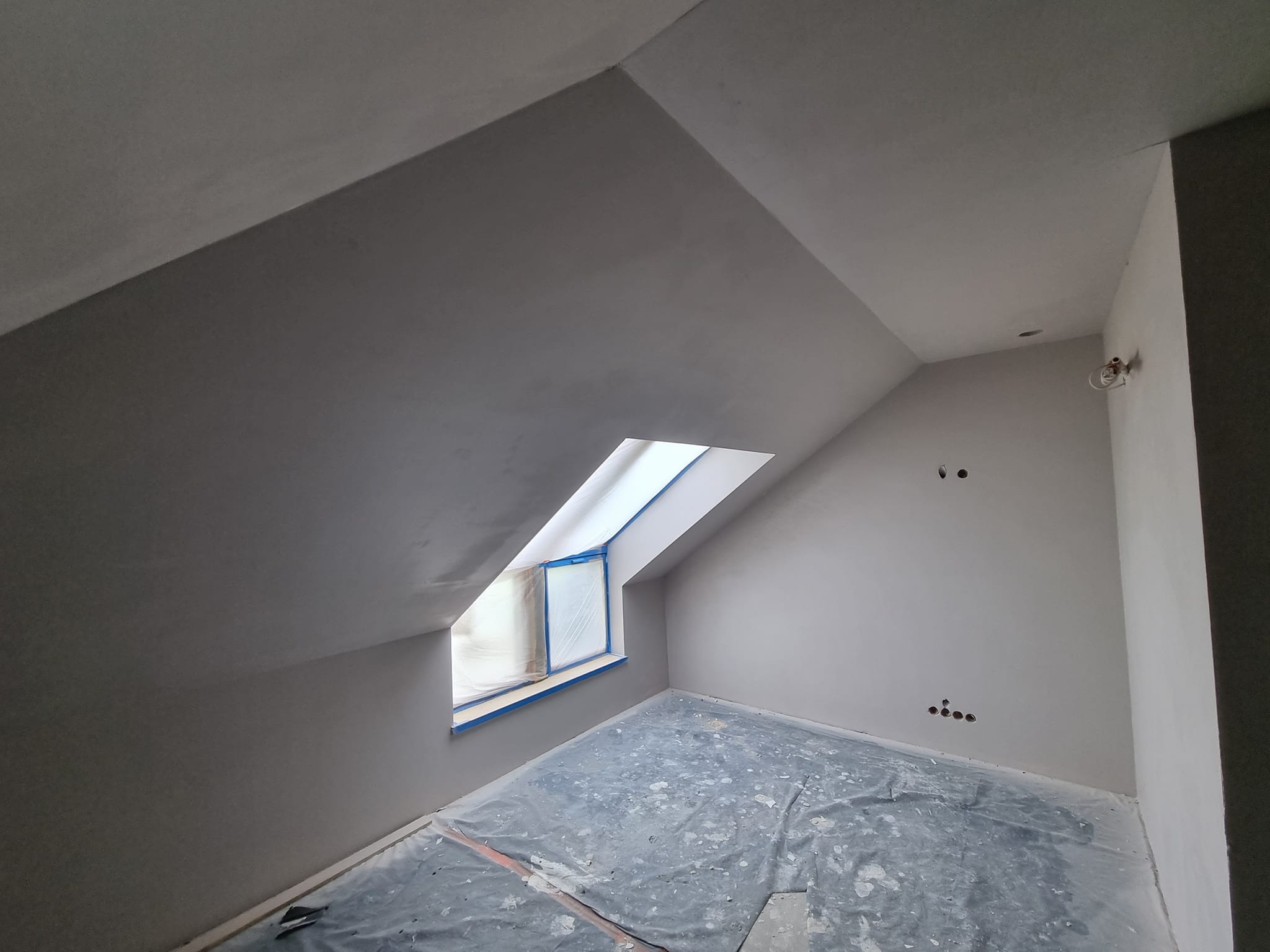 Pokój na poddaszu po szpachlowaniu, przygotowany do malowania. Okno dachowe zabezpieczone folią, podłoga przykryta. Widoczne gładkie ściany i sufit.