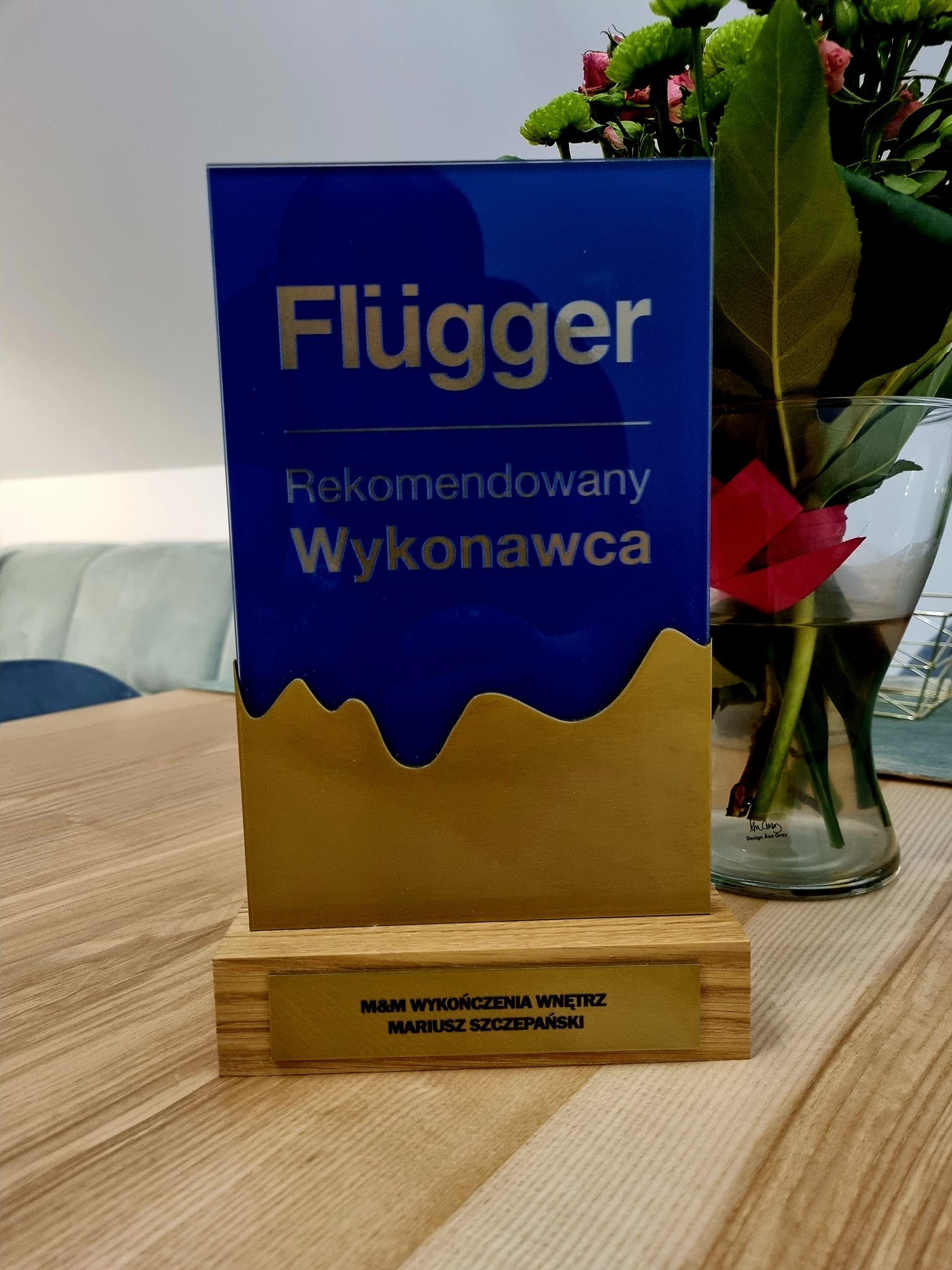 Nagroda 'Rekomendowany Wykonawca Flügger' dla M&M Wykończenia Wnętrz Mariusza Szczepańskiego, stojąca na drewnianym stole obok wazonu z kwiatami.