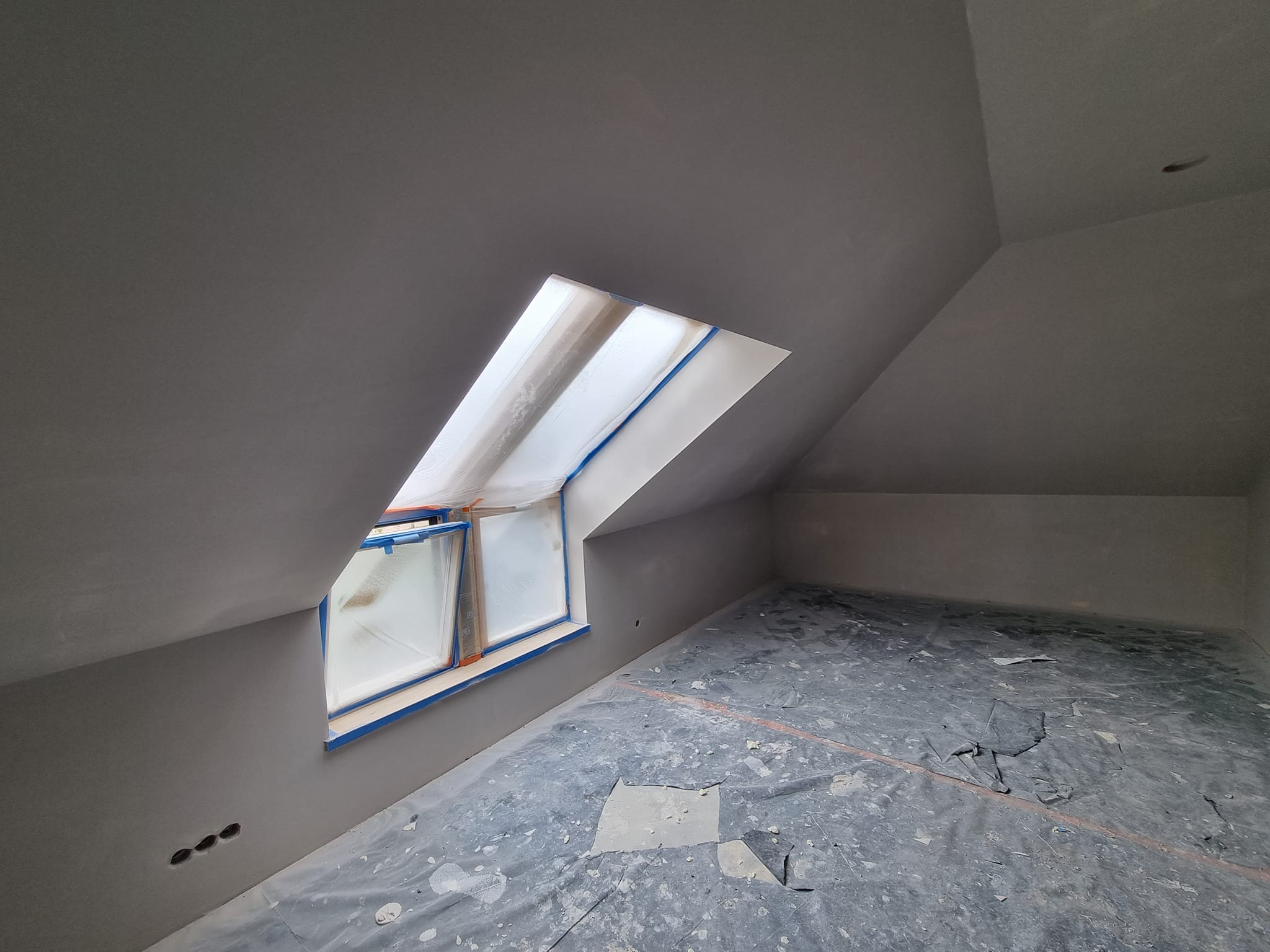 Pokój na poddaszu po szpachlowaniu, okno zabezpieczone folią, podłoga przykryta plandeką. Widoczne gładkie ściany i przygotowanie do dalszych prac wykończeniowych.