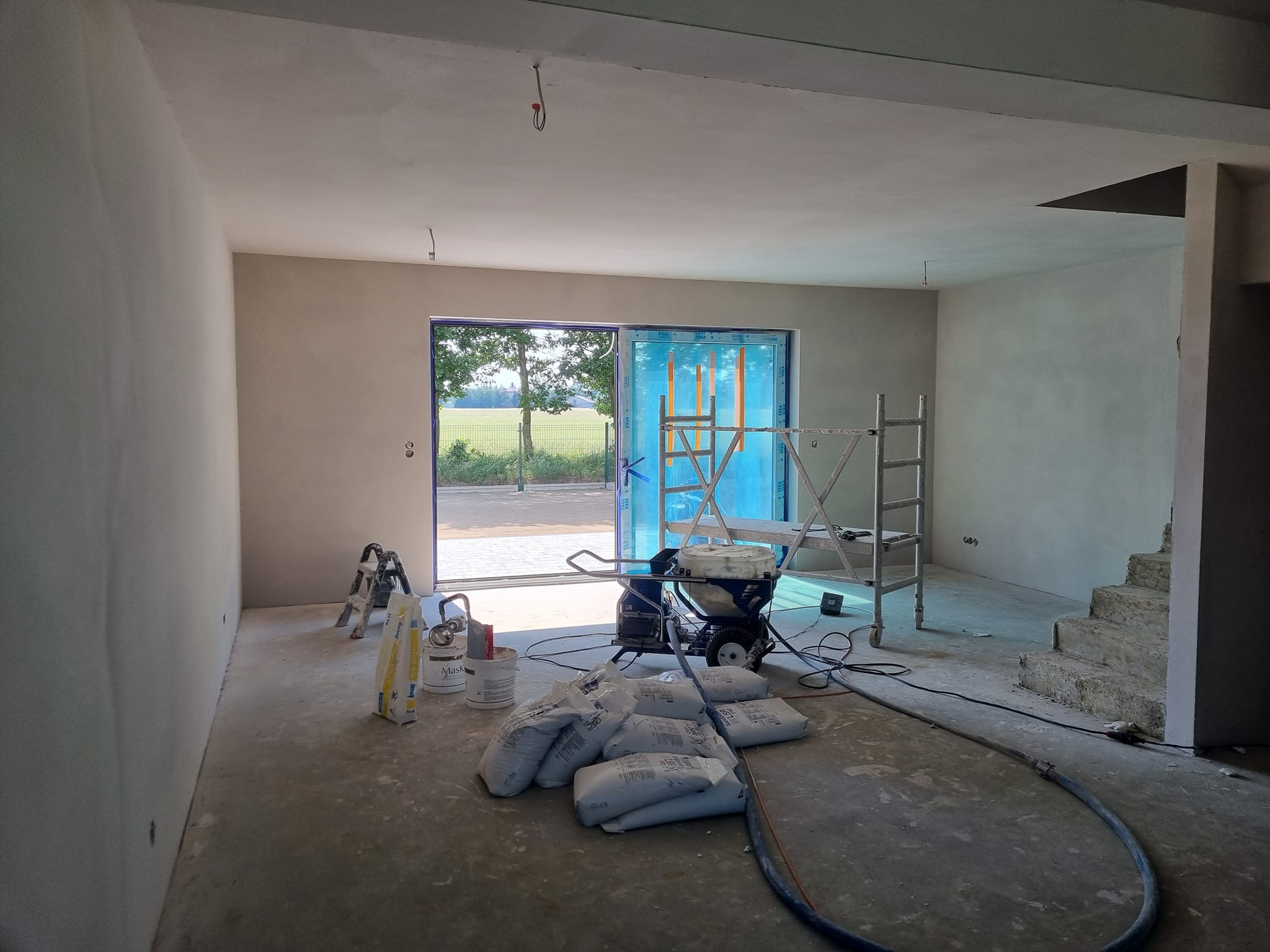 Wnętrze w trakcie prac: ściany po szpachlowaniu, maszyna do natrysku, worki z materiałem, drabina. Widok na zewnątrz przez otwarte drzwi balkonowe.