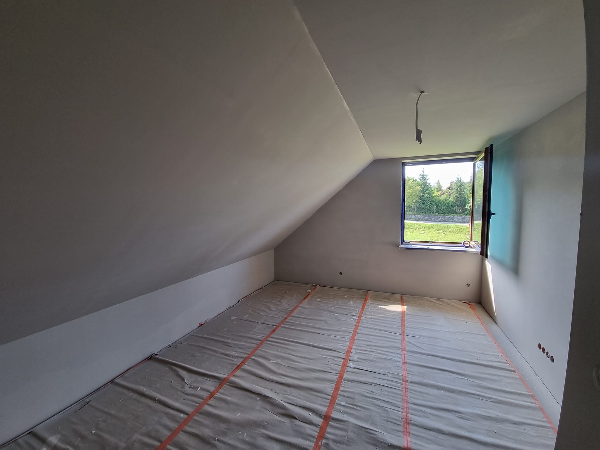 Pokój na poddaszu po szpachlowaniu ścian i sufitu, zabezpieczony folią malarską, z oknem wychodzącym na zieleń. Widoczne gniazdka elektryczne.