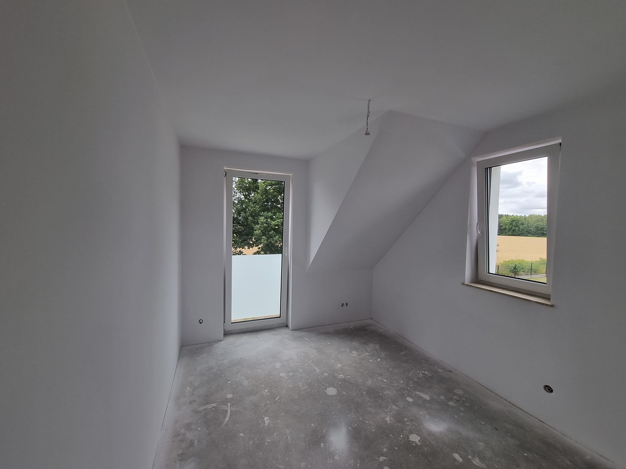 Wnętrze pokoju na poddaszu po szpachlowaniu i malowaniu na biało. Widoczne okno z widokiem na zieleń i pole, surowa podłoga, gładkie ściany. Przygotowanie do dalszych prac wykończeniowych.
