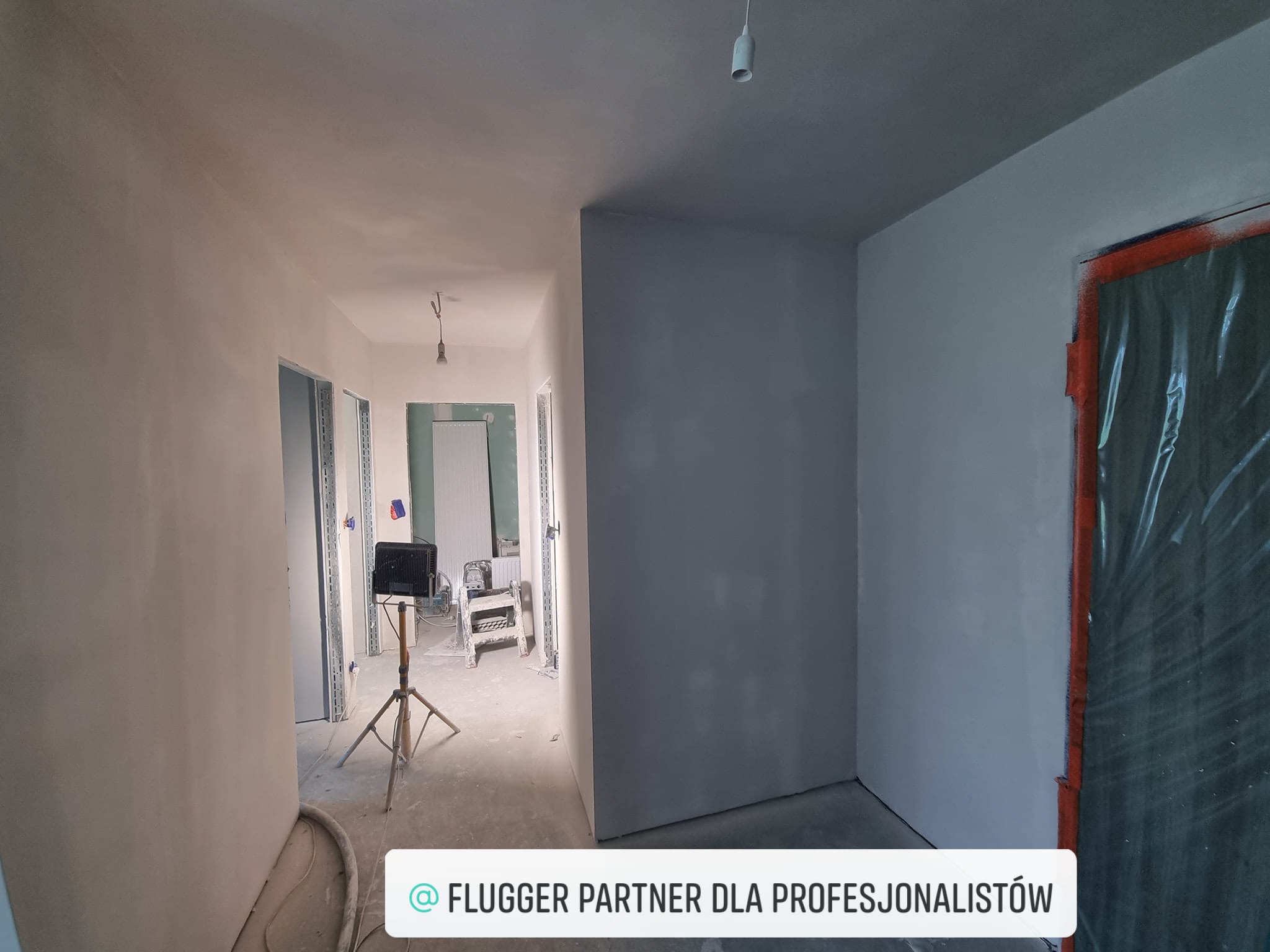 Wnętrze po szpachlowaniu: gładkie, szare ściany w korytarzu, zabezpieczone drzwi folią. Widoczny sprzęt budowlany i oświetlenie. Etap przygotowania do malowania.