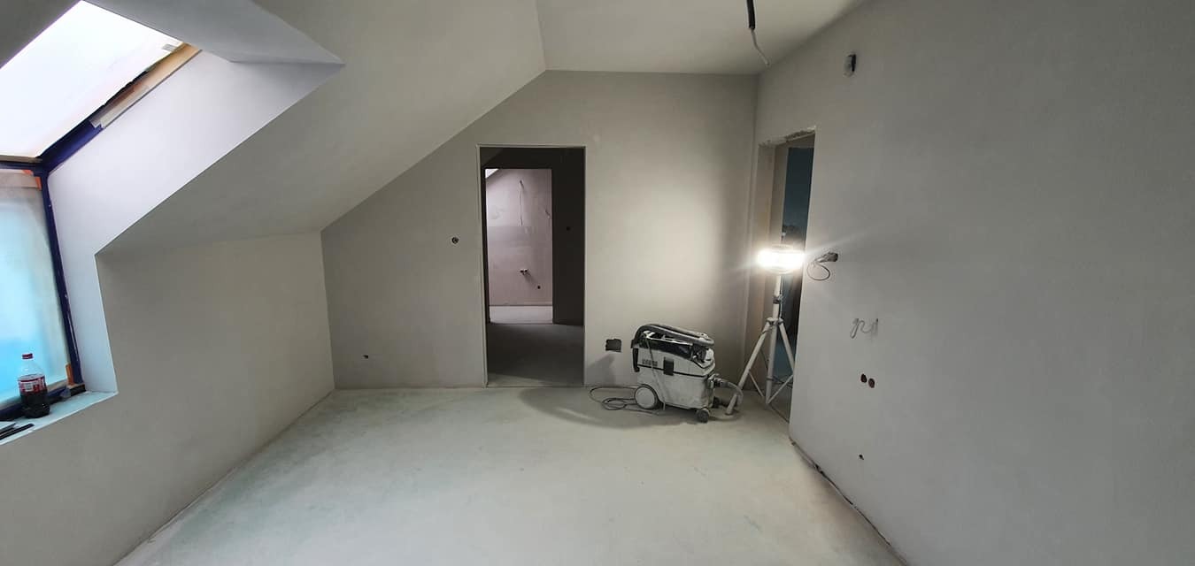 Pokój na poddaszu po szpachlowaniu i malowaniu na biało. Widoczny świetlik, drzwi, lampa na statywie i odkurzacz przemysłowy.