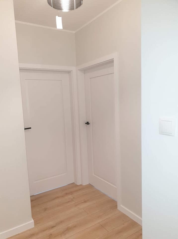 Jasny korytarz po remoncie: białe ściany, drewnopodobna podłoga, dwie białe drzwi z czarnymi klamkami, nowoczesna lampa sufitowa i włącznik światła.