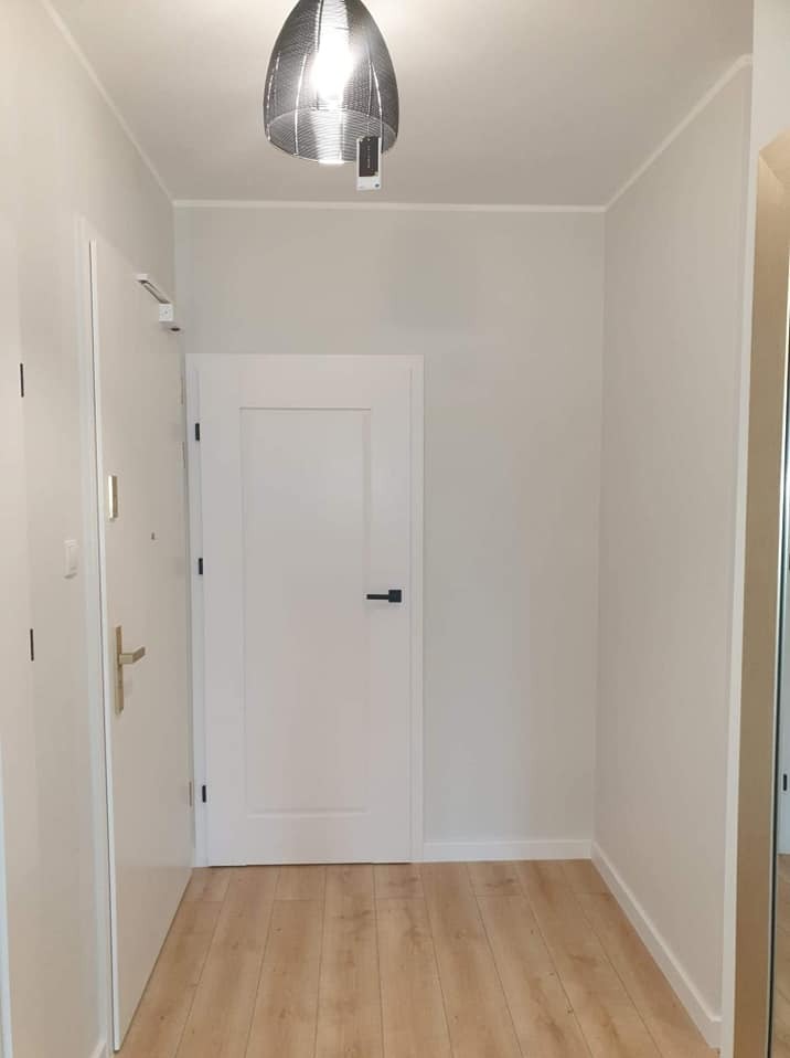 Wnętrze po odświeżeniu: Jasny korytarz z drewnopodobną podłogą, białe ściany i drzwi z czarną klamką. Designerska lampa wisząca dodaje nowoczesnego charakteru.
