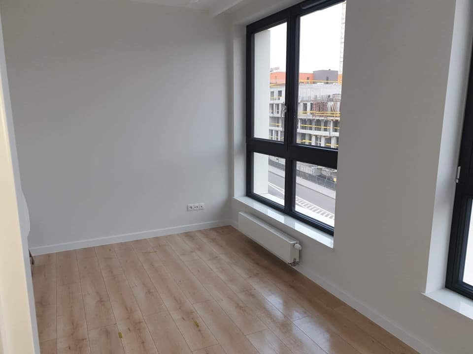 Pomalowane na biało wnętrze pokoju z drewnianą podłogą i czarnymi oknami z widokiem na budowę. Świeżo wykończone ściany, listwy przypodłogowe i grzejnik.