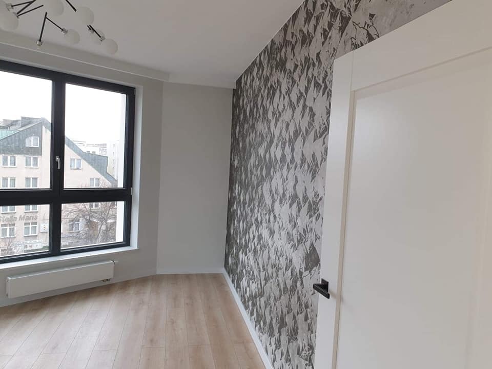 Pokój po remoncie: ściana z tapetą w geometryczny wzór, jasna podłoga, okno z widokiem na miasto, białe drzwi z czarną klamką. Nowoczesne oświetlenie sufitowe.