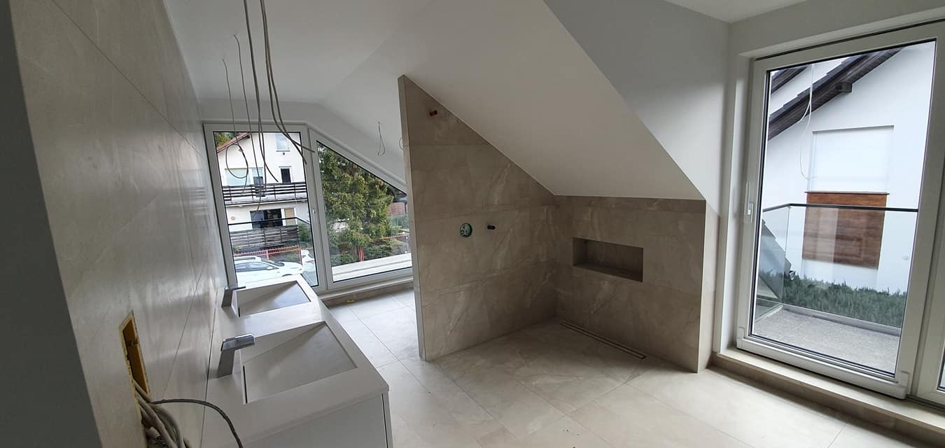 Wykończona łazienka na poddaszu z oknami, beżowe płytki na ścianach i podłodze, biała umywalka z dwiema misami. Widok na zewnątrz przez okna.