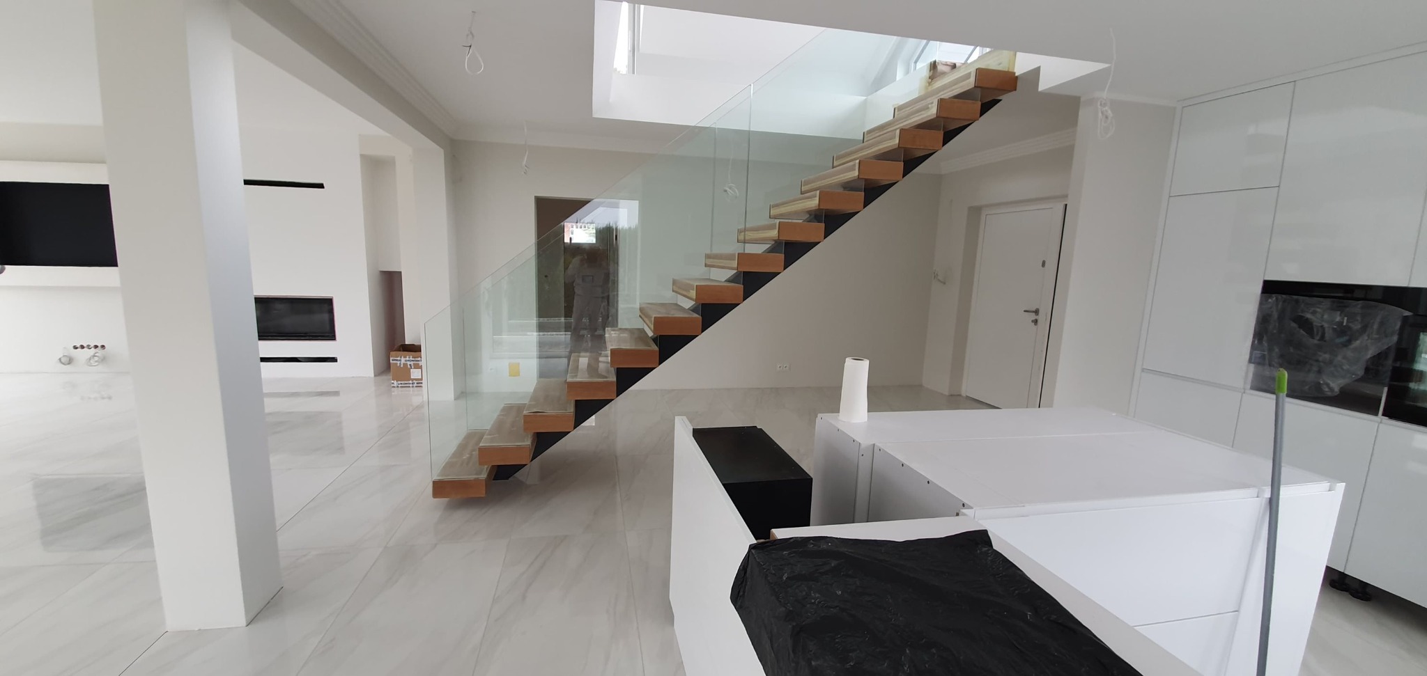 Nowoczesne wnętrze: białe ściany, marmurowe podłogi, drewniane schody z szklaną balustradą. Widoczne elementy zabudowy kuchennej i minimalistyczny kominek.