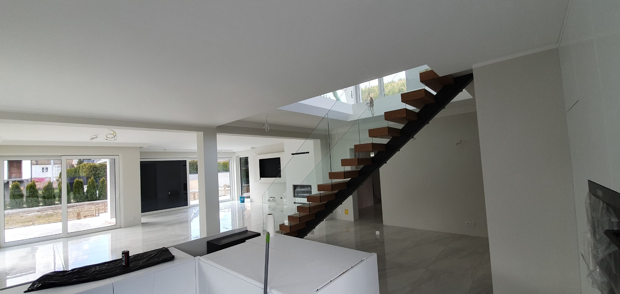 Jasne, przestronne wnętrze domu po remoncie: białe ściany, marmurowe podłogi, nowoczesne schody z drewnianymi stopniami i szklaną balustradą. Widok na ogród przez duże okna.