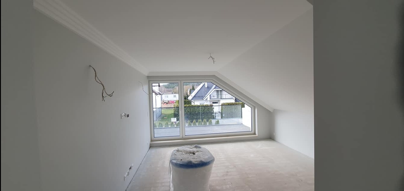 Pokój na poddaszu po malowaniu na biało, z oknem dachowym i listwami ozdobnymi. Widoczne przewody elektryczne i zabezpieczony materiał budowlany.