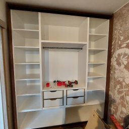 Jacek Bojarski - Wnętrze szafy wnękowej z białymi półkami i dwiema szufladami, widoczne narzędzia montażowe, brązowe obramowanie, tapeta w kwiaty w tle.