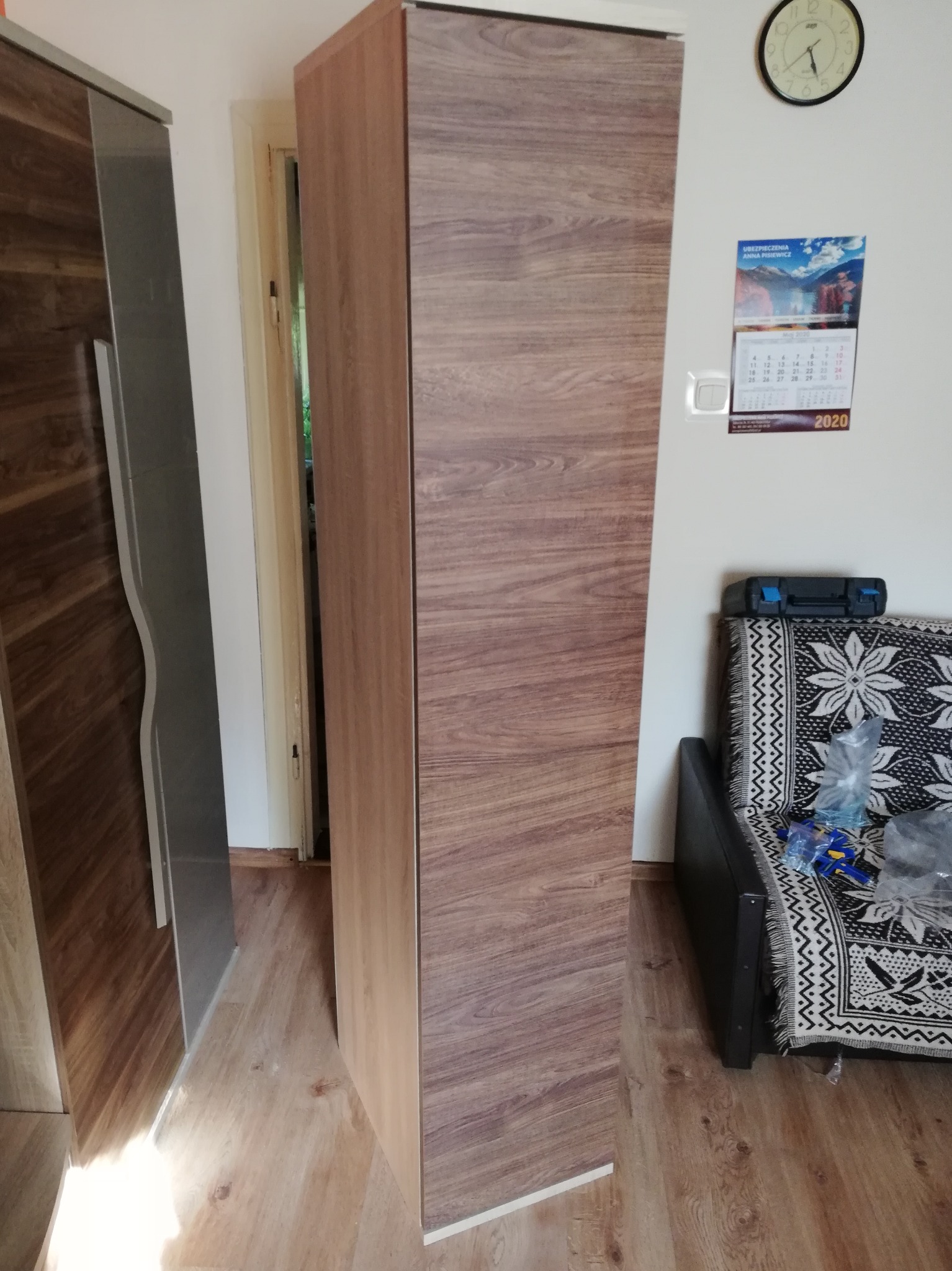 Wysoka szafa z okleiną imitującą drewno, ustawiona w rogu pokoju. Widoczny fragment drugiej szafy po lewej stronie, zegar i kalendarz na ścianie w tle.