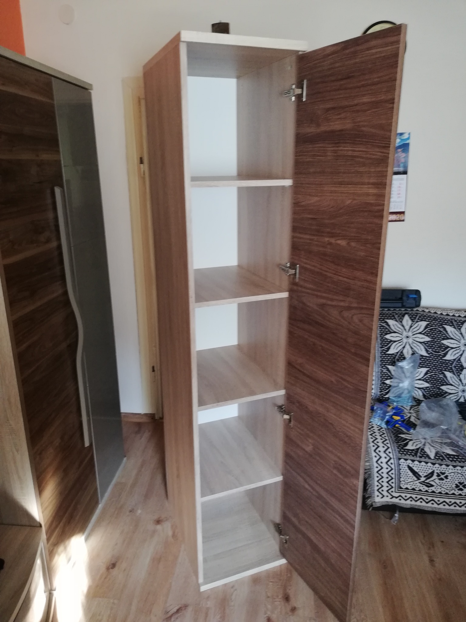 Otwarta szafa z półkami w odcieniach drewna i bieli. Widoczne zawiasy i wnętrze szafy. Obok stoi inna szafa z przesuwnymi drzwiami. W tle kanapa z wzorem kwiatowym.