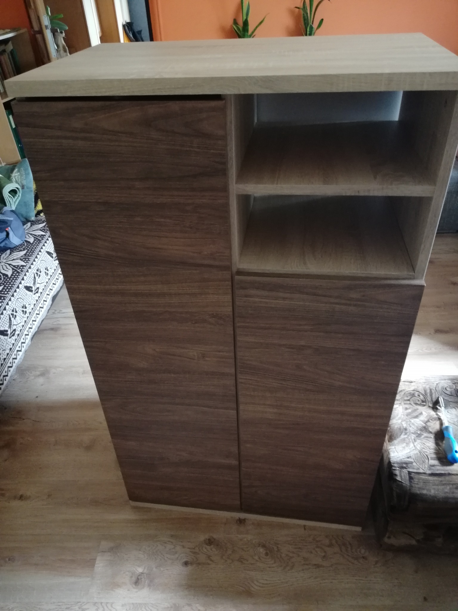 Nowoczesna szafka z dwiema szafkami zamykanymi i otwartymi półkami, wykonana z drewna w odcieniach brązu i beżu, na tle podłogi w panelach.