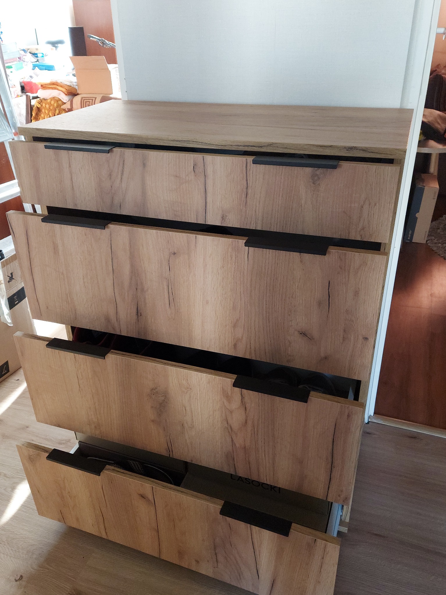 Czterodrzwiowa komoda z drewnopodobnymi frontami i czarnymi uchwytami, częściowo wysunięte szuflady ukazują zawartość. Widoczny fragment podłogi.