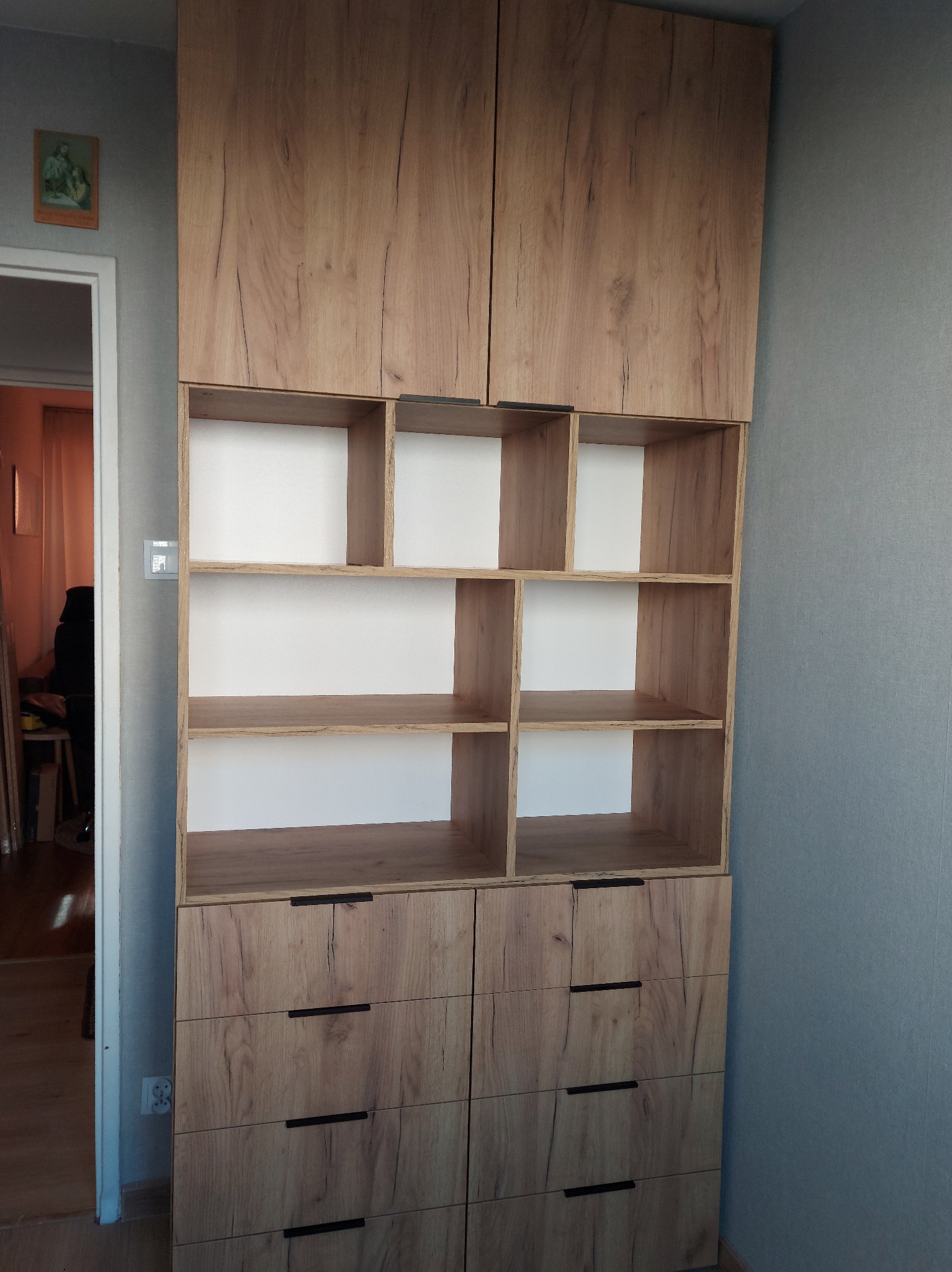 Drewniana szafa na wymiar z szufladami i półkami, z widoczną strukturą drewna, ustawiona przy szarej ścianie, z obrazkiem religijnym nad drzwiami.