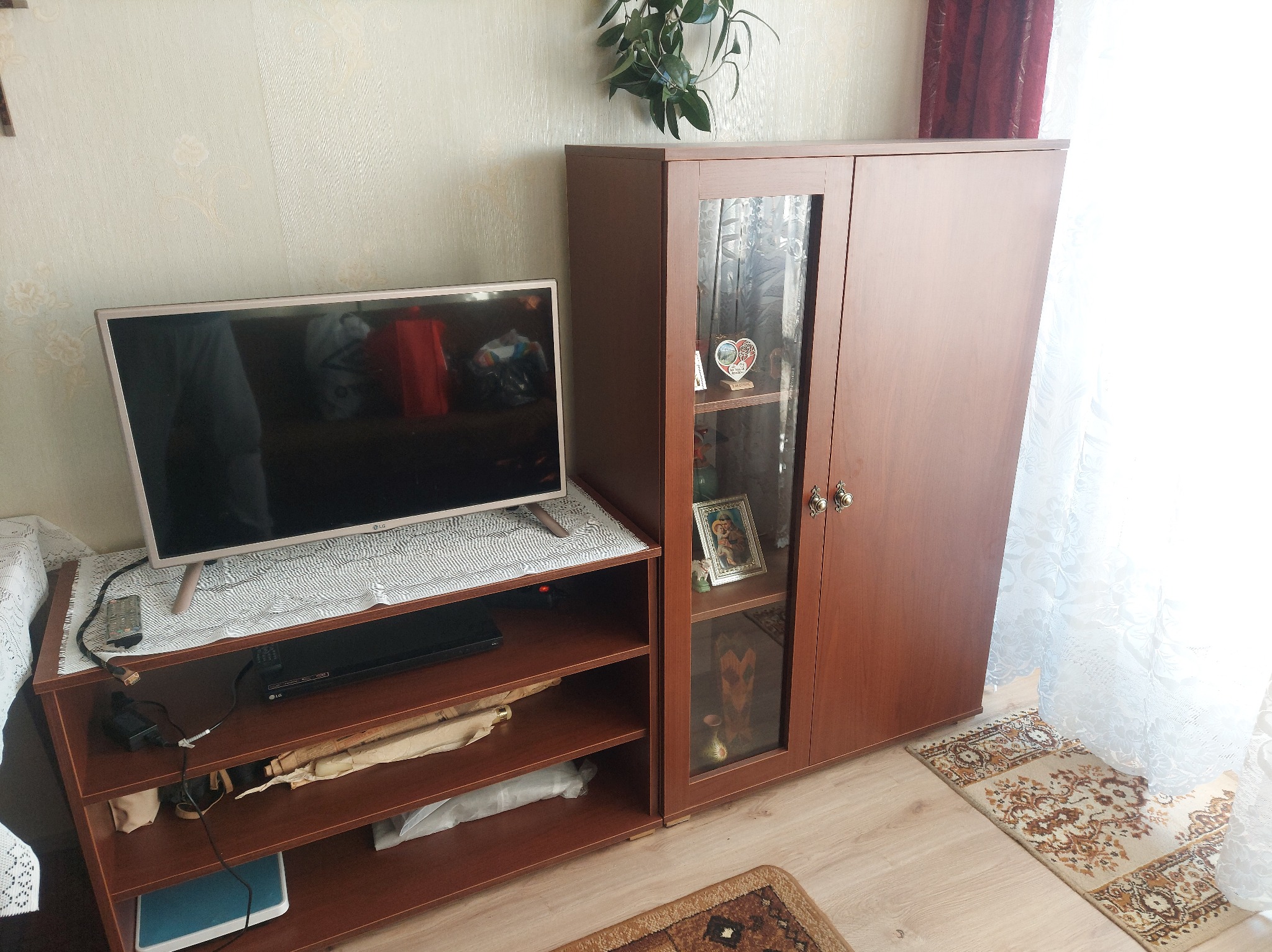 Brązowa szafka z przeszklonymi drzwiami i półkami, obok szafka RTV z telewizorem. Na półkach akcesoria, na szafce serweta. W tle firanka i roślina.