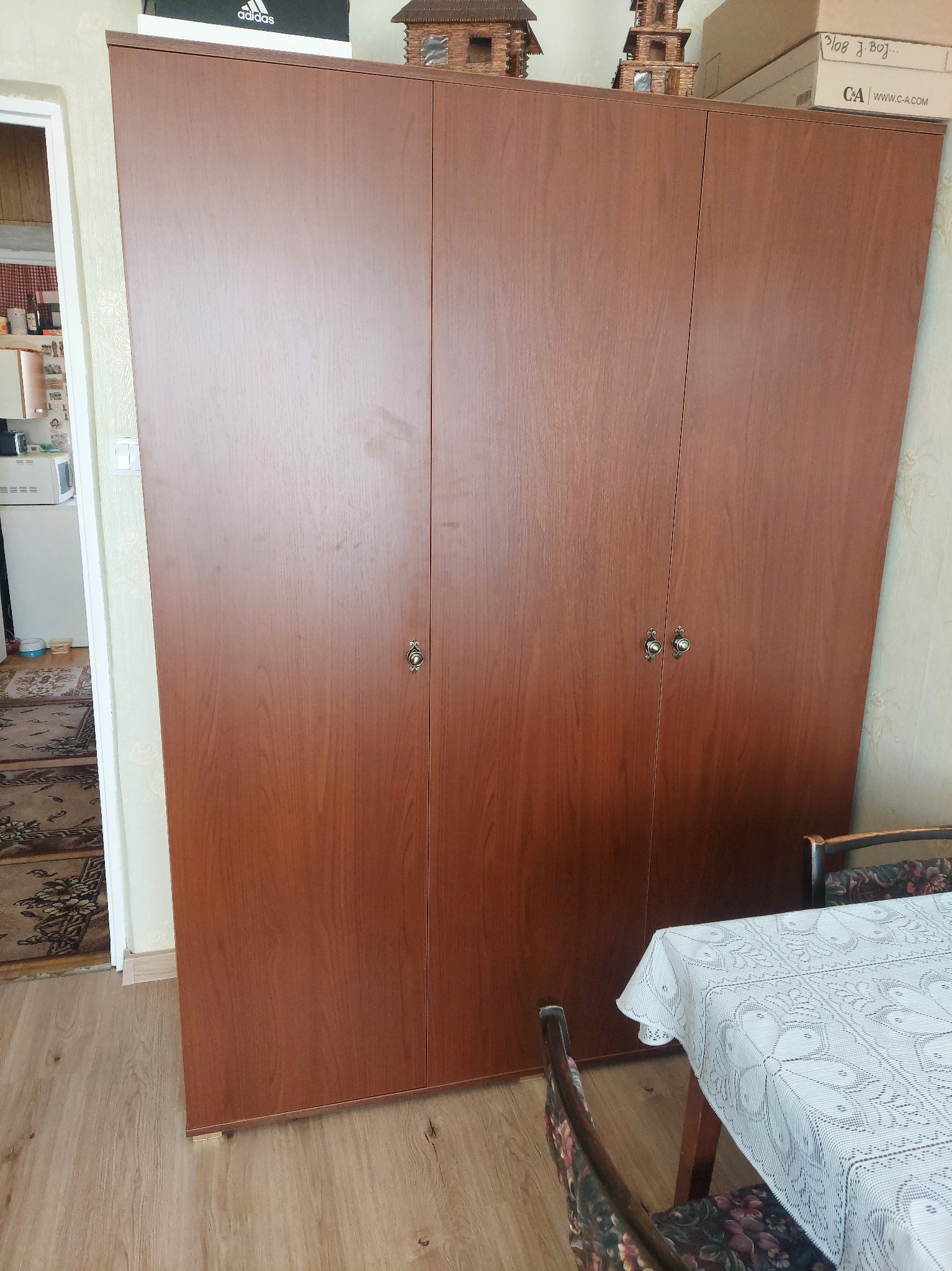 Trzydrzwiowa szafa z okleiną w odcieniu drewna, z ozdobnymi uchwytami, częściowo zasłonięta stołem z koronkowym obrusem. Na szafie pudełka i dekoracyjne domki.